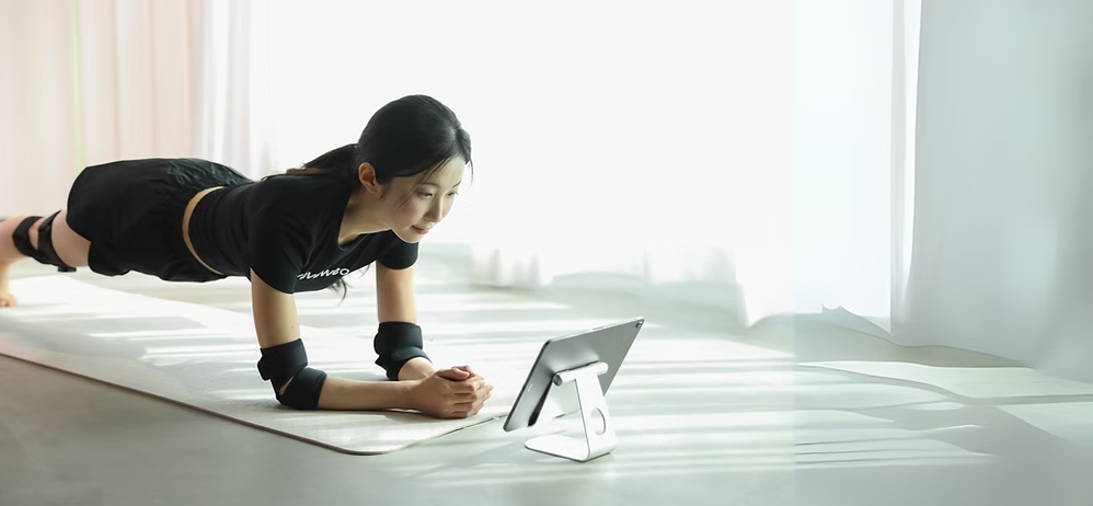 A woman doing a plank exercise on a yoga mat while looking at a tablet on a stand in a bright room.