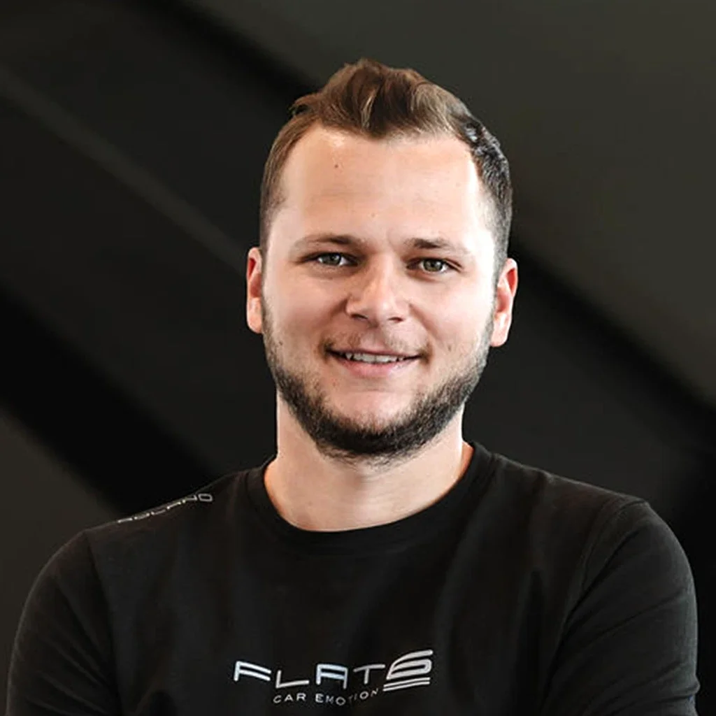 Ein junger Mann mit kurzen braunen Haaren und Bart, trägt ein schwarzes T-Shirt mit der Aufschrift 'FLAT6 CAR EMOTION'.