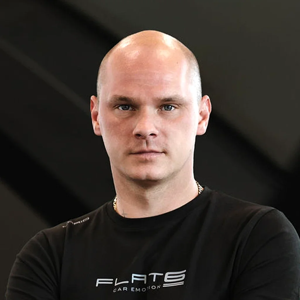 Porträt eines Mannes mit Glatze, schwarzem T-Shirt mit weißem Schriftzug, in einem dunklen Hintergrund.