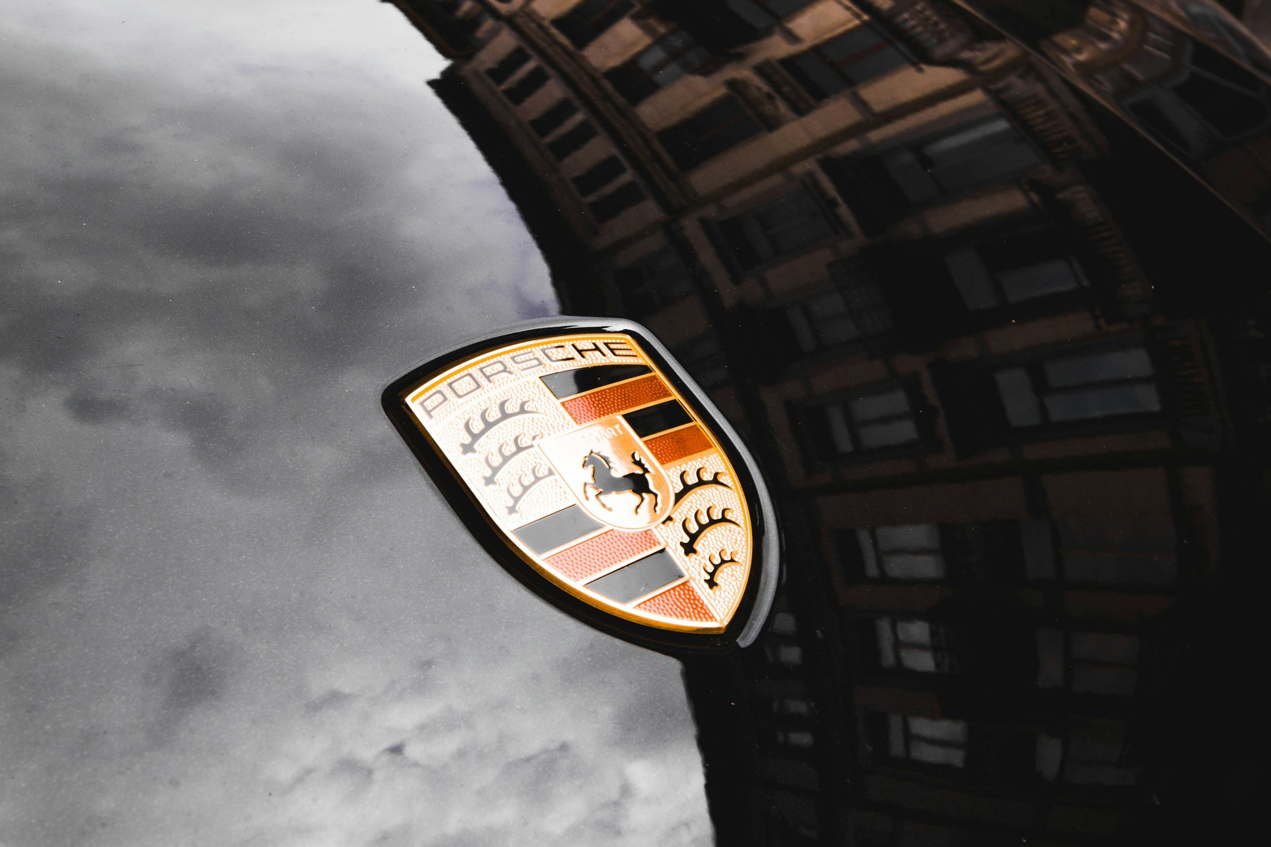 Das Porsche-Logo auf einem schwarzen Auto, mit einer reflektierenden sturmverhangenen Stadtlandschaft im Hintergrund.