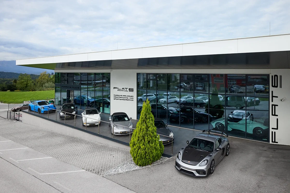 FLAT6 in Elixhausen. Ein modernes Autohaus mit großen Glasscheiben, in dem mehrere Porsche und Performance-Fahrzeuge ausgestellt sind. Vor dem Gebäude sind weitere Autos parkiert.