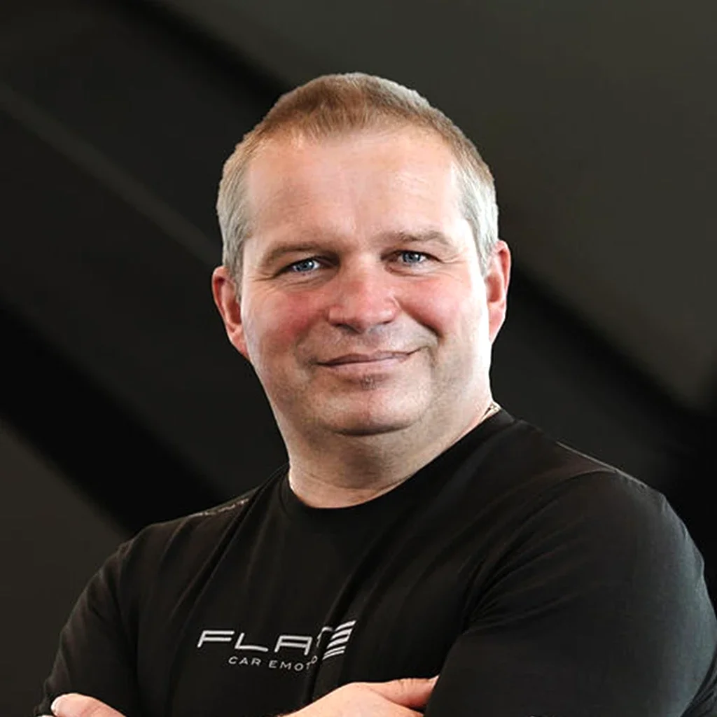 Ein Mann mit kurzen Haaren, blauer Augen und einem schwarzen T-Shirt, der vor einem dunklen Hintergrund steht.