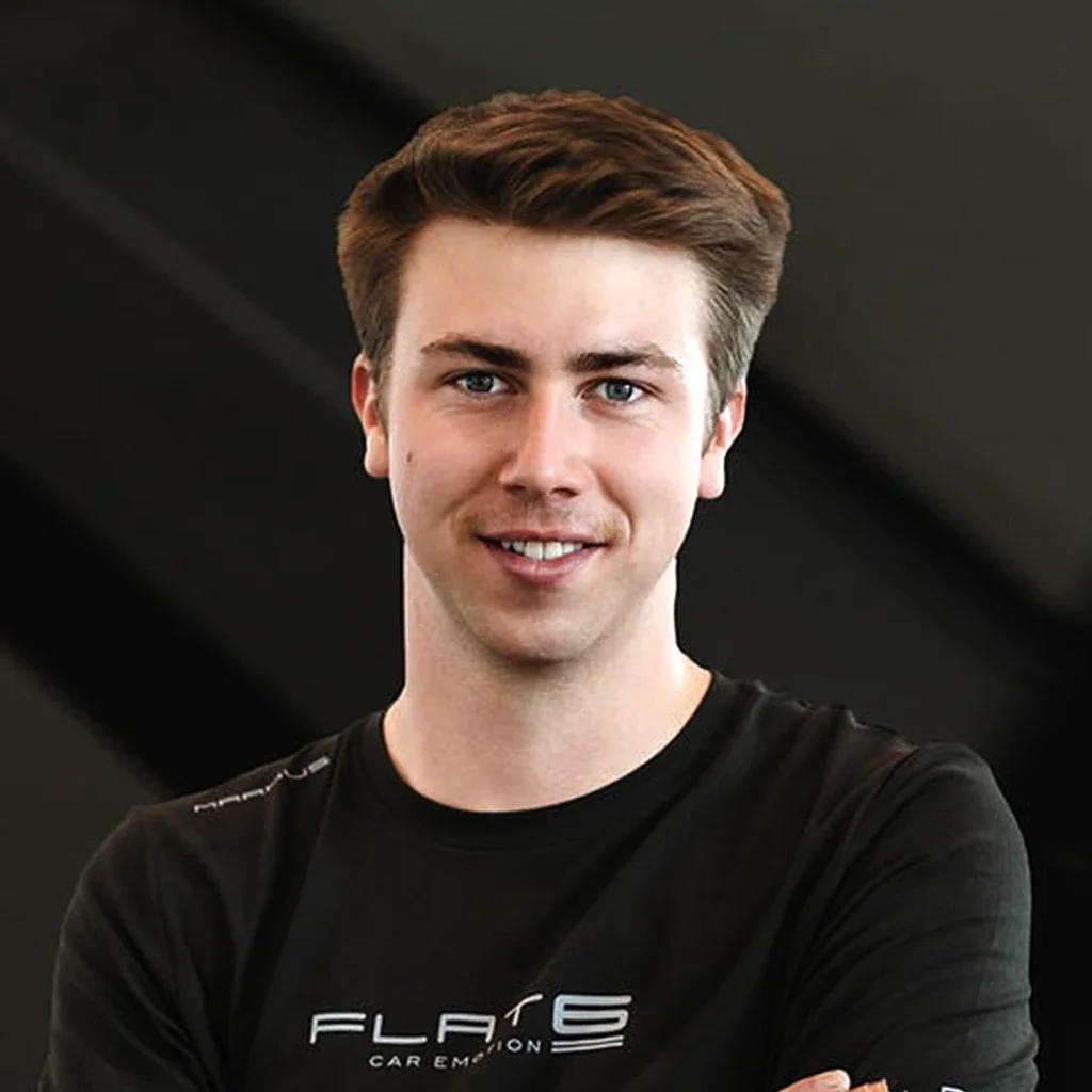Ein junger Mann mit braunem Haar und blauen Augen, trägt ein schwarzes T-Shirt mit weißem Druck, steht vor einem dunklen Hintergrund.