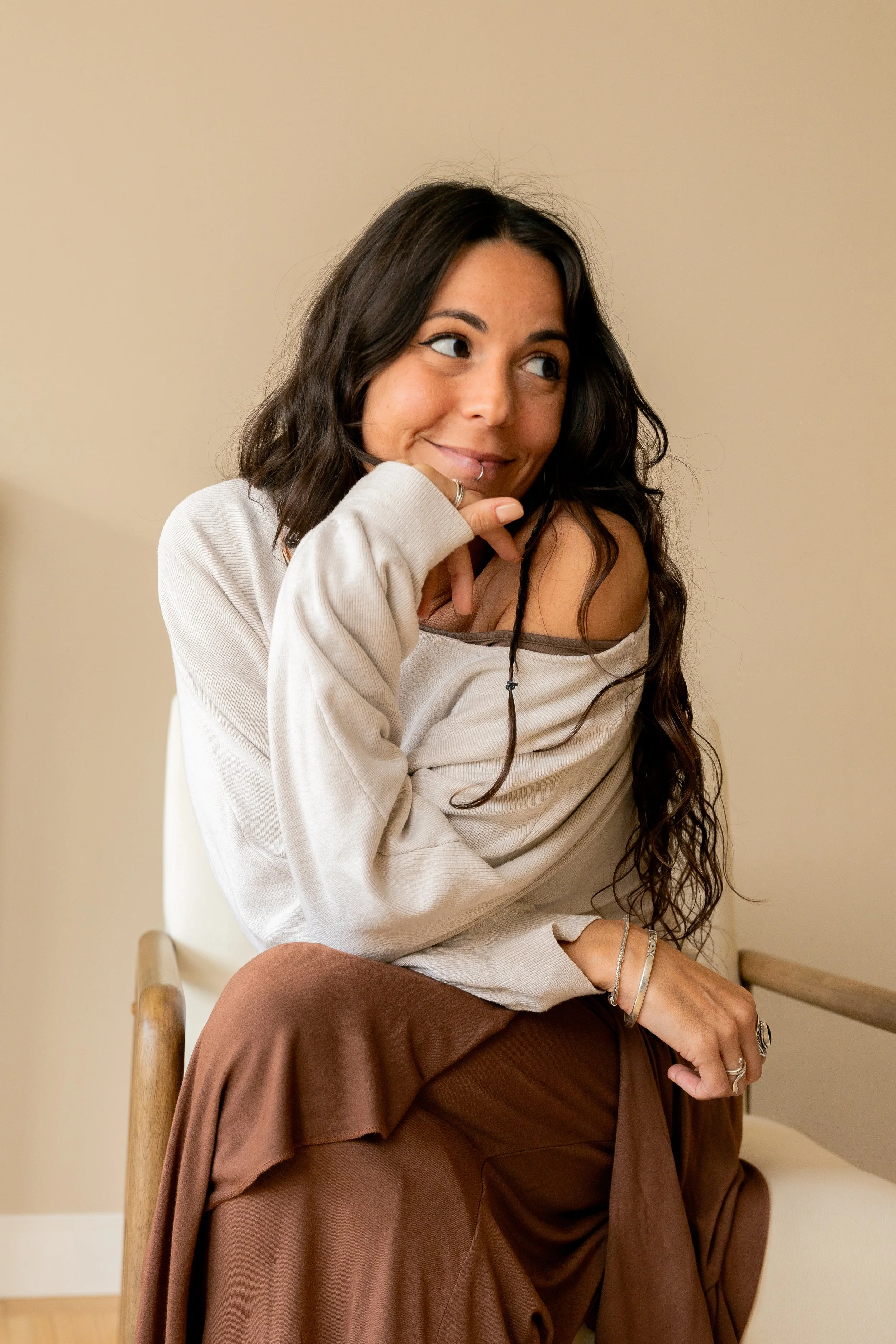 Una mujer con cabello rizado y oscuro, sonriendo y sosteniéndose la barbilla con su mano izquierda, vestida con un suéter beige y una falda marrón, sentada en una silla en un ambiente interior.