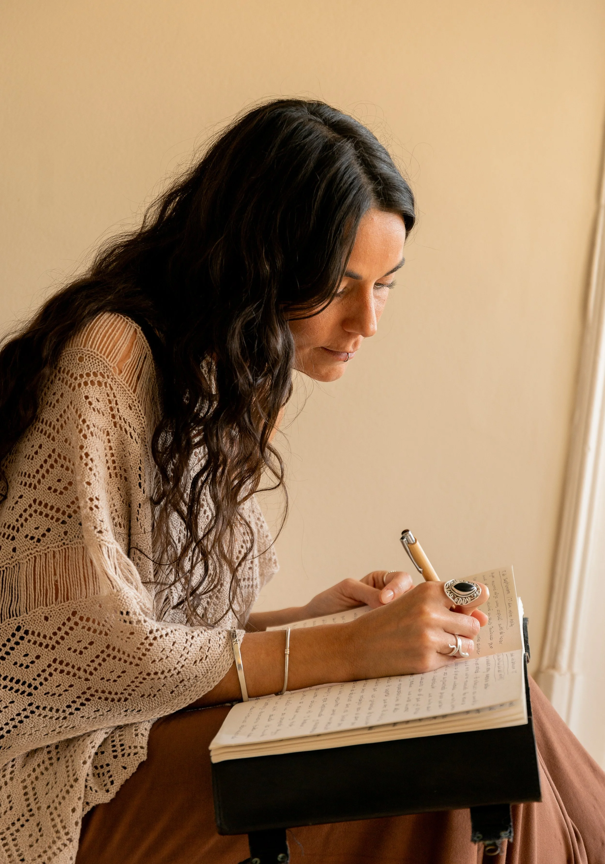 Mujer con cabello rizado oscuro, usando un suéter de crochet beige, escribiendo en un cuaderno sobre una mesa.
