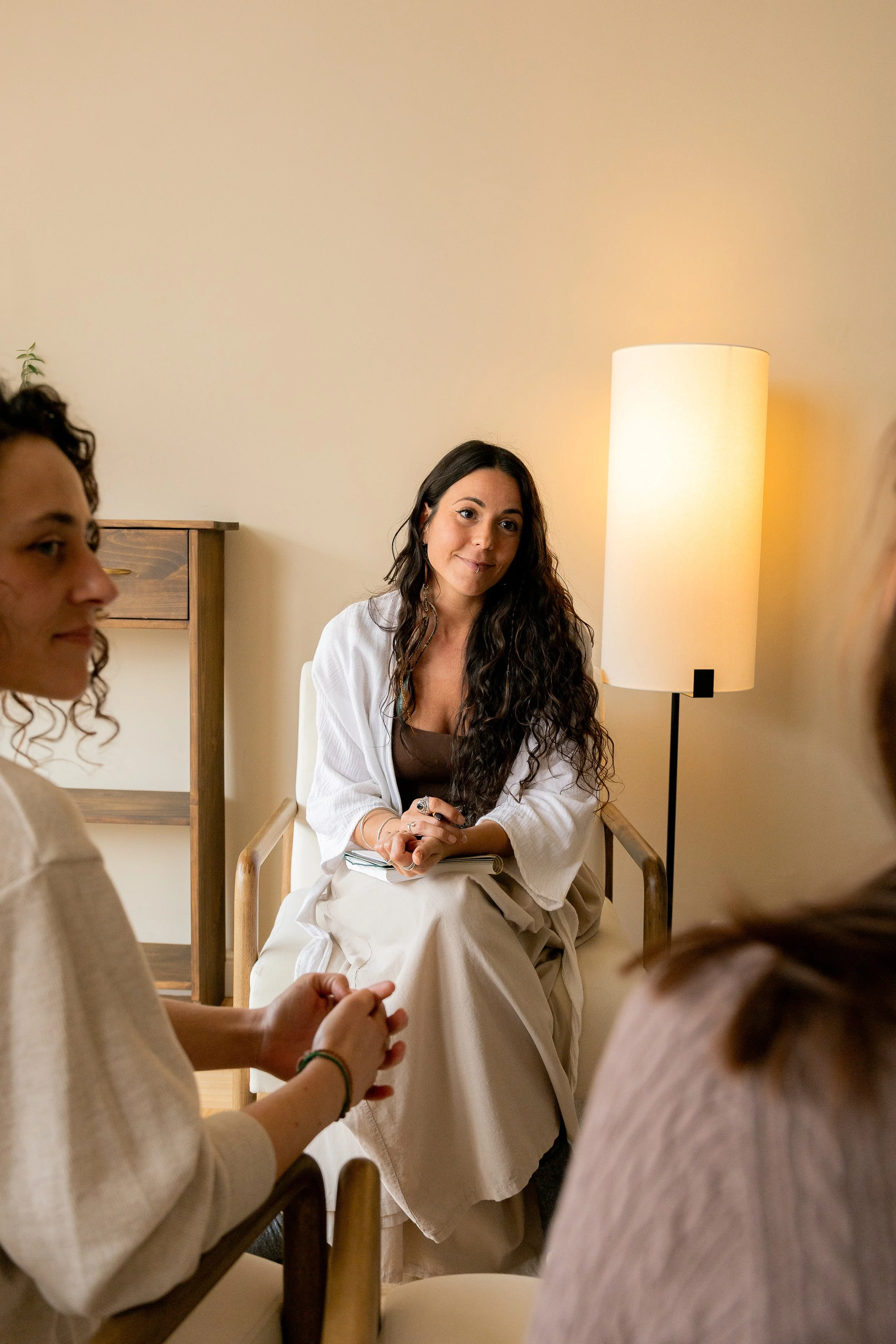 Grupo de personas en una sala, una mujer con cabello rizado oscuro, vestida con ropa blanca, sentada en una silla y sosteniendo un cuaderno, escucha a otras dos mujeres que están de espaldas, en un ambiente cálido con lámpara de pie de luz suave.