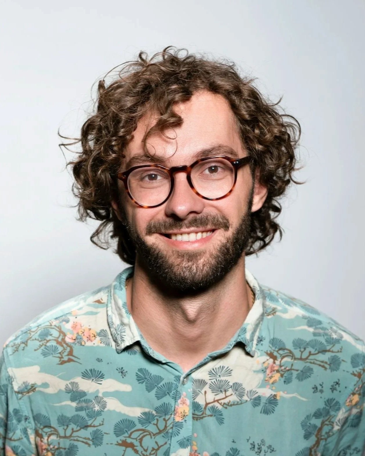 Ung mand med brunt, krøllet hår, briller, skæg og smilende, iført hawaii skjorte mod en hvid baggrund.