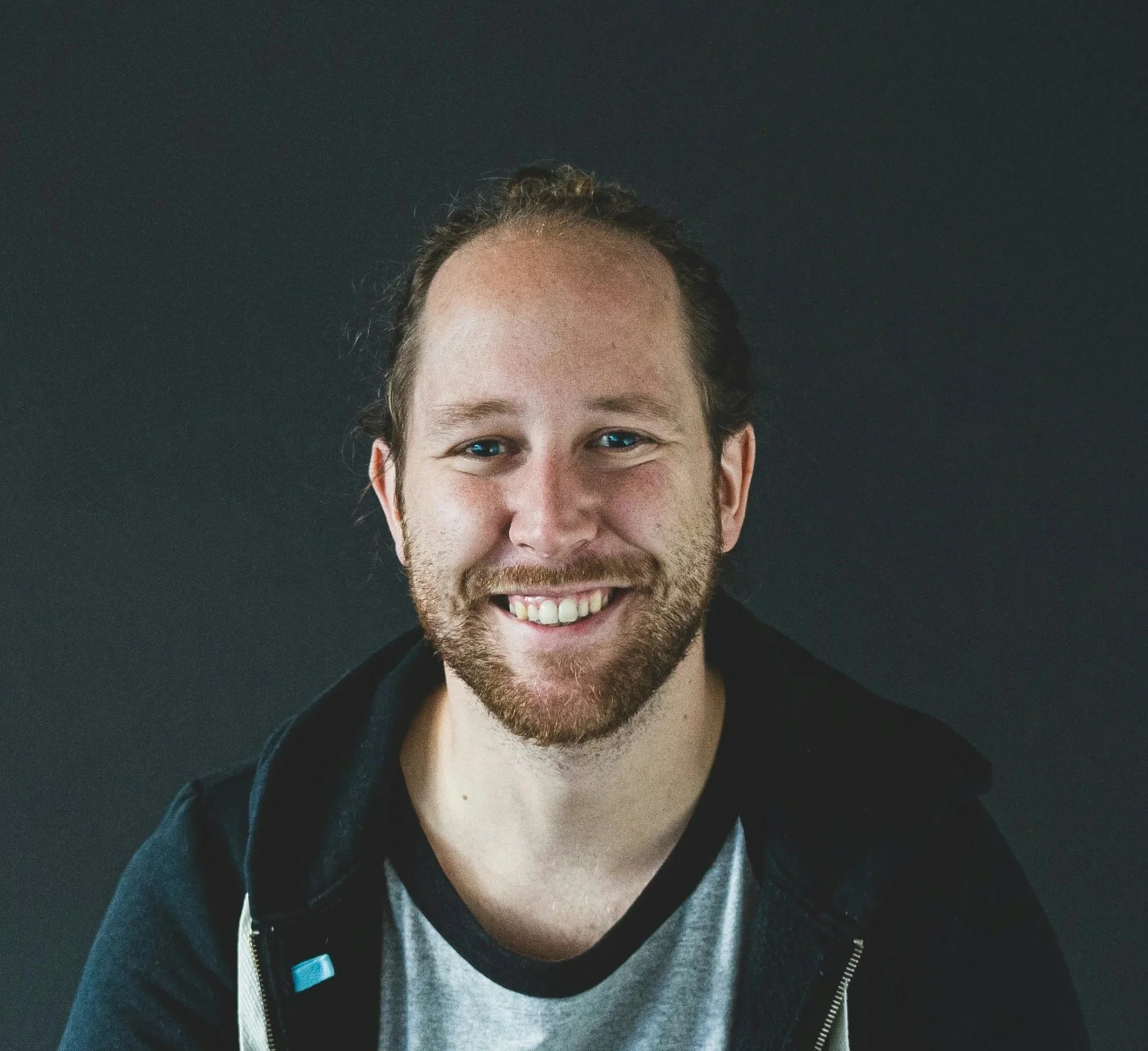 Portræt af en smilende mand med skæg og brunt hår, iført grå T-shirt og sort jakke, mod en mørk baggrund.