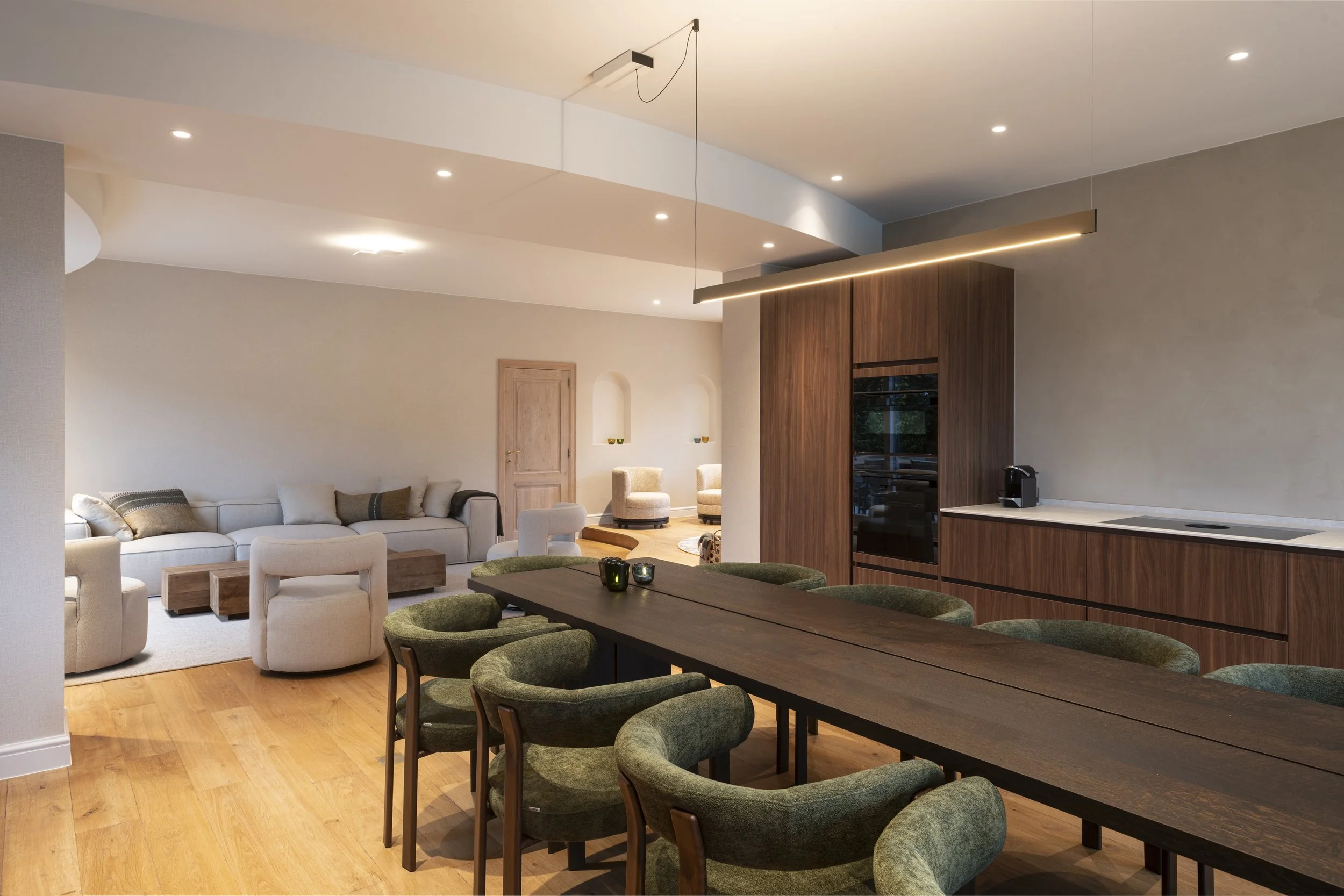 Modern open-concept living and dining room with wood flooring, a large light-colored sofa, green upholstered chairs around a dining table, and a sleek kitchen with wood cabinetry and a built-in oven.