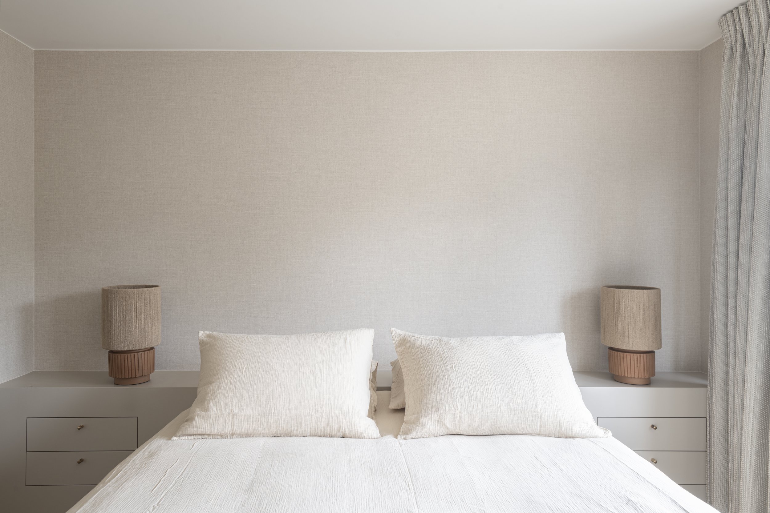 Minimalist bedroom with a white bed, two beige bedside tables with matching lamps, a plain beige wall, and a curtain on the right side.