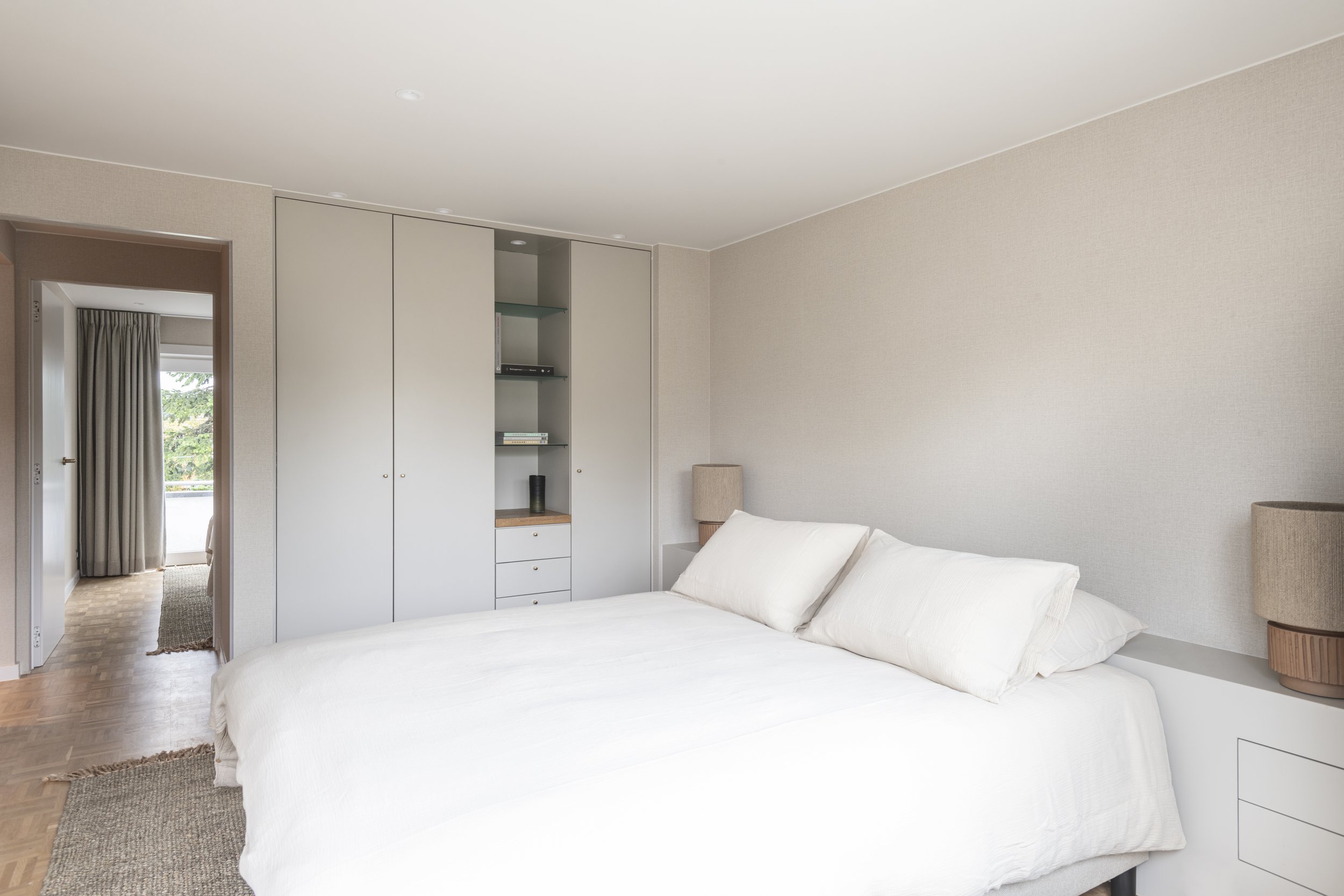 A bedroom with a white bed, beige lamps, a built-in wardrobe, and a small open shelf with books and decor, with a doorway leading to a room with a window and curtains.