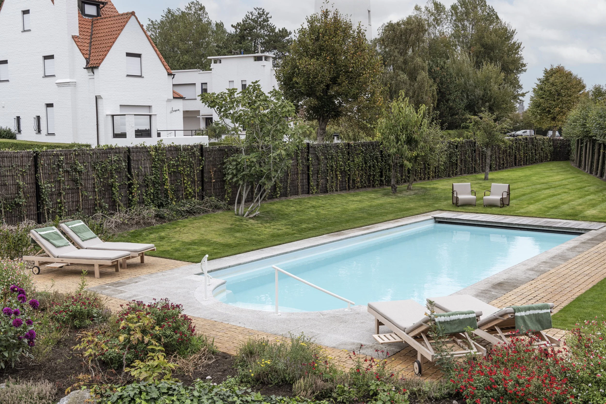 A backyard with a swimming pool, surrounded by green grass and garden beds with colorful flowers. Two lounge chairs with green towels are near the pool. In the background, there is a tall wooden fence, several trees, and a white modern house with a r