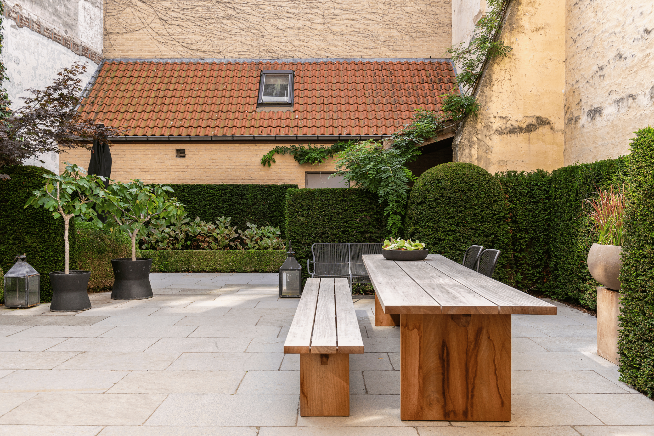 Outdoor patio with a long wooden table and chairs, surrounded by potted plants, greenery, and brick walls.