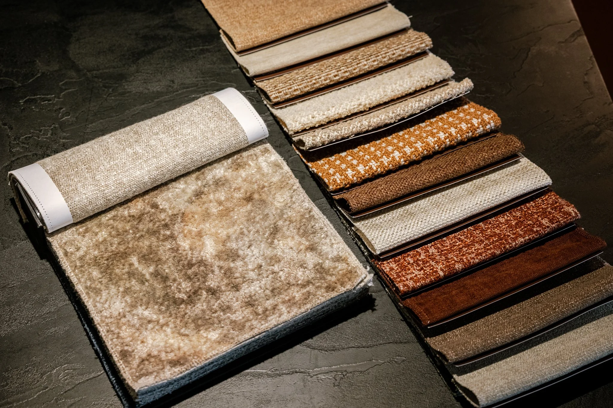 A display of rolled and flat fabric swatches in various shades of beige, brown, and orange, laid out on a dark surface.