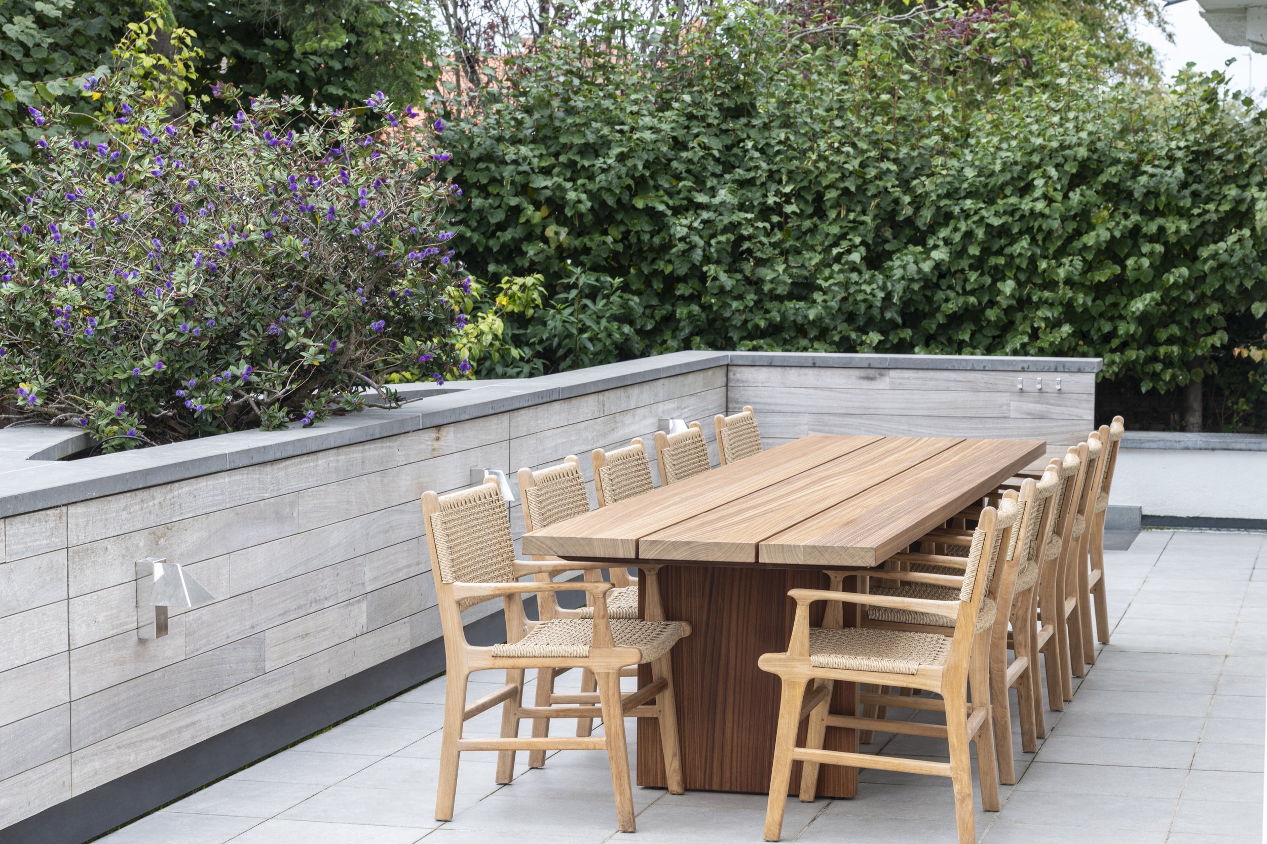 A long wooden outdoor dining table with matching chairs, set against a backdrop of green hedges and flowering bushes, with a concrete wall and tiled patio floor.