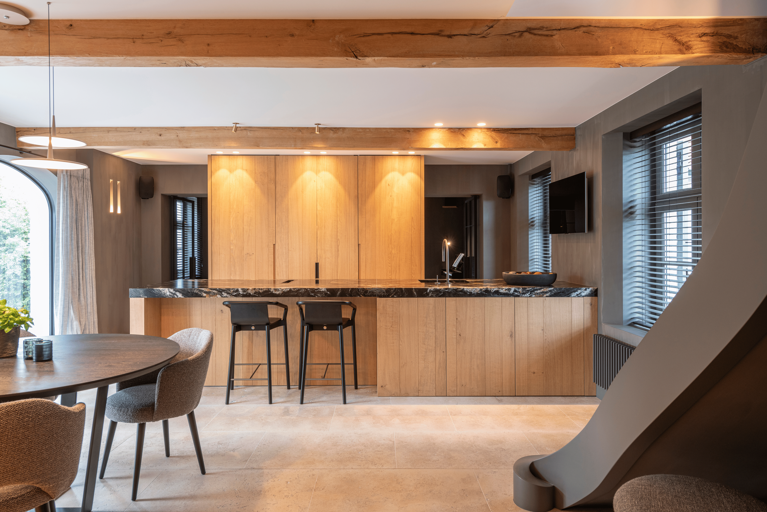 Interior of a modern kitchen with wooden cabinets, a marble countertop island, black bar stools, and large windows with blinds.