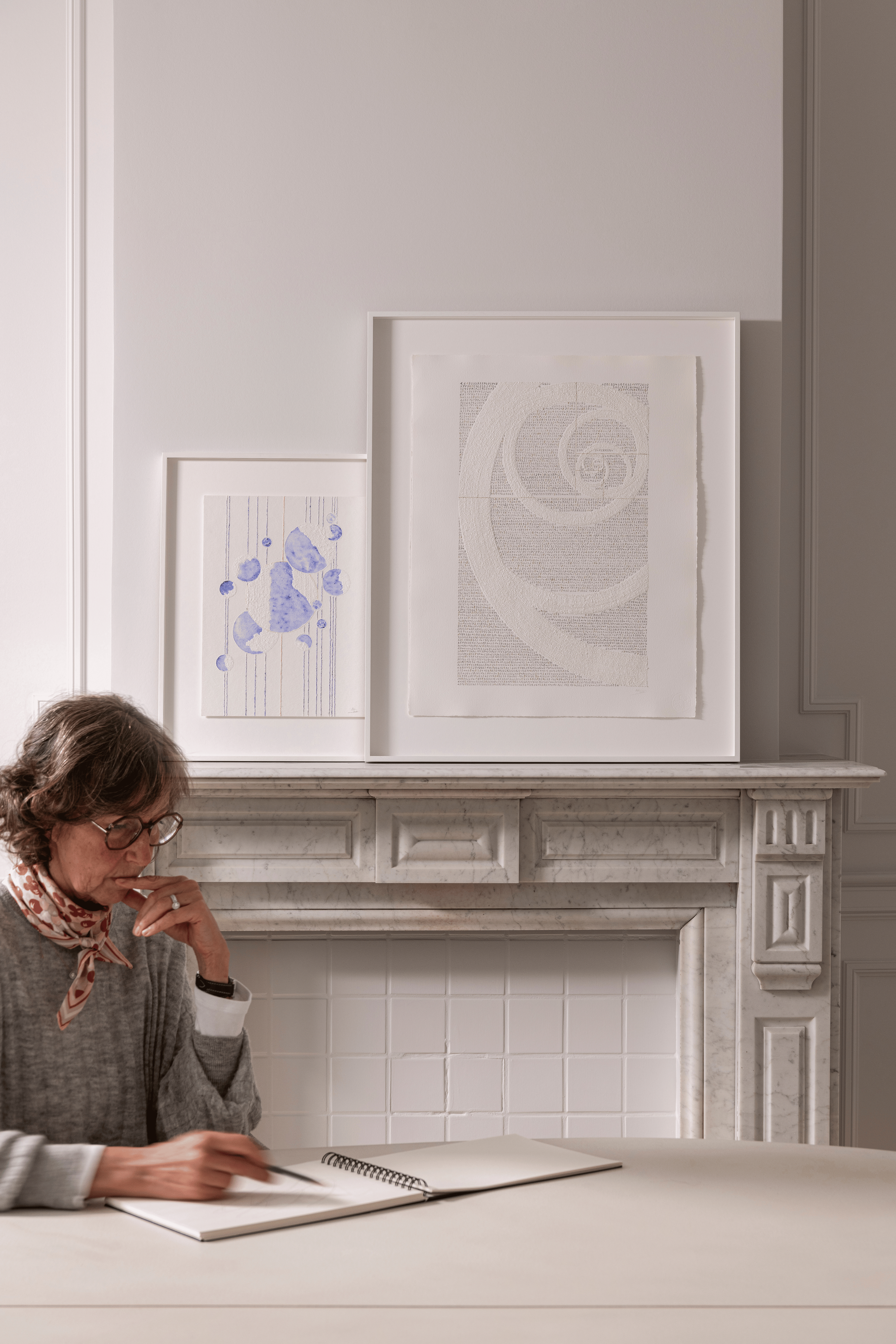 A woman with glasses and a scarf around her neck sitting at a table, writing in a notebook, in a room with a white marble fireplace and artwork on the wall.