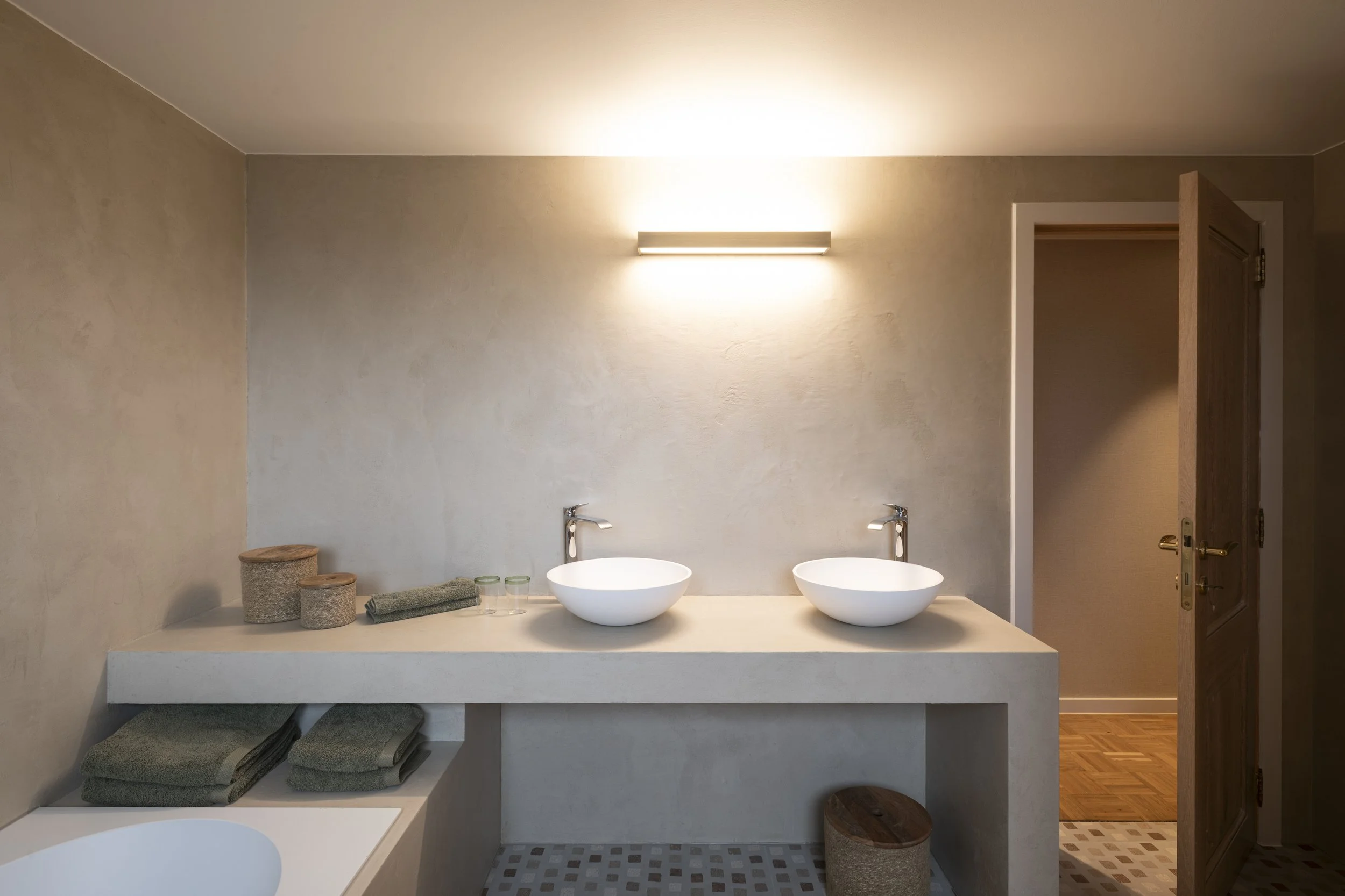A bathroom vanity with two vessel sinks, gray towels, and storage baskets on a white countertop, with a wall-mounted light fixture above.
