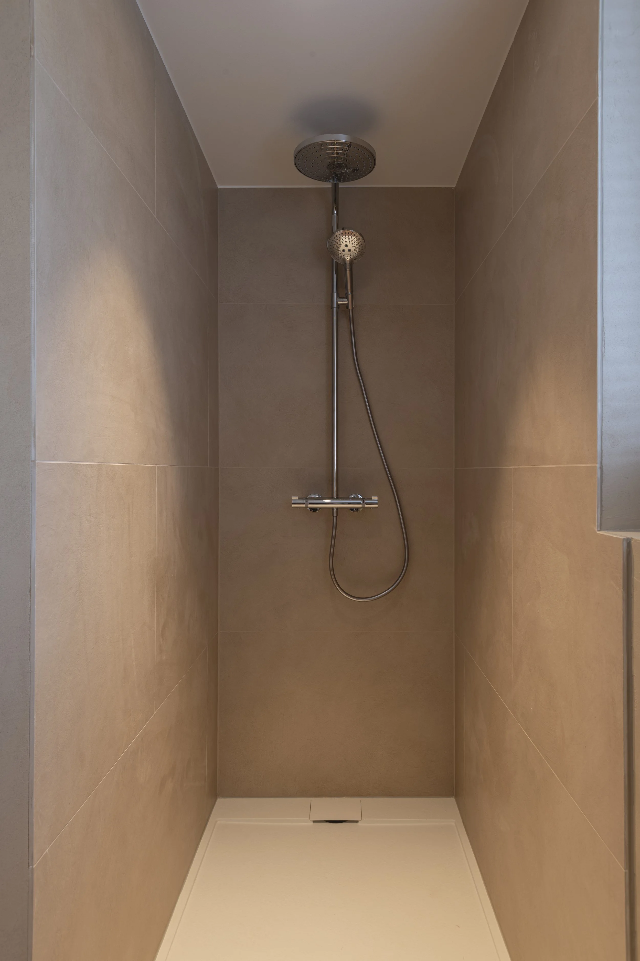 A plain shower with beige tiled walls, a silver showerhead, a handheld shower attachment, and a white floor drain.