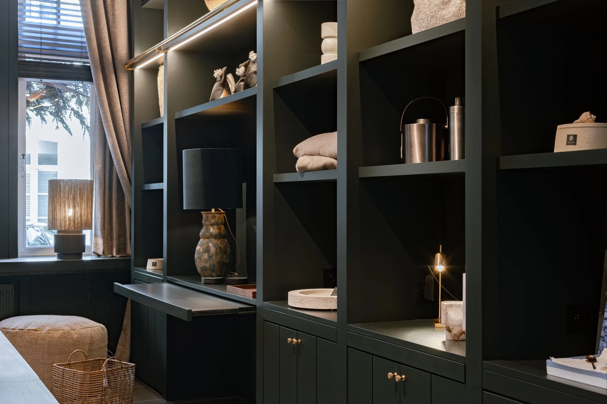 Dark green cabinet with decorative items and lighting, next to a window with curtains and a lamp in a cozy interior space.