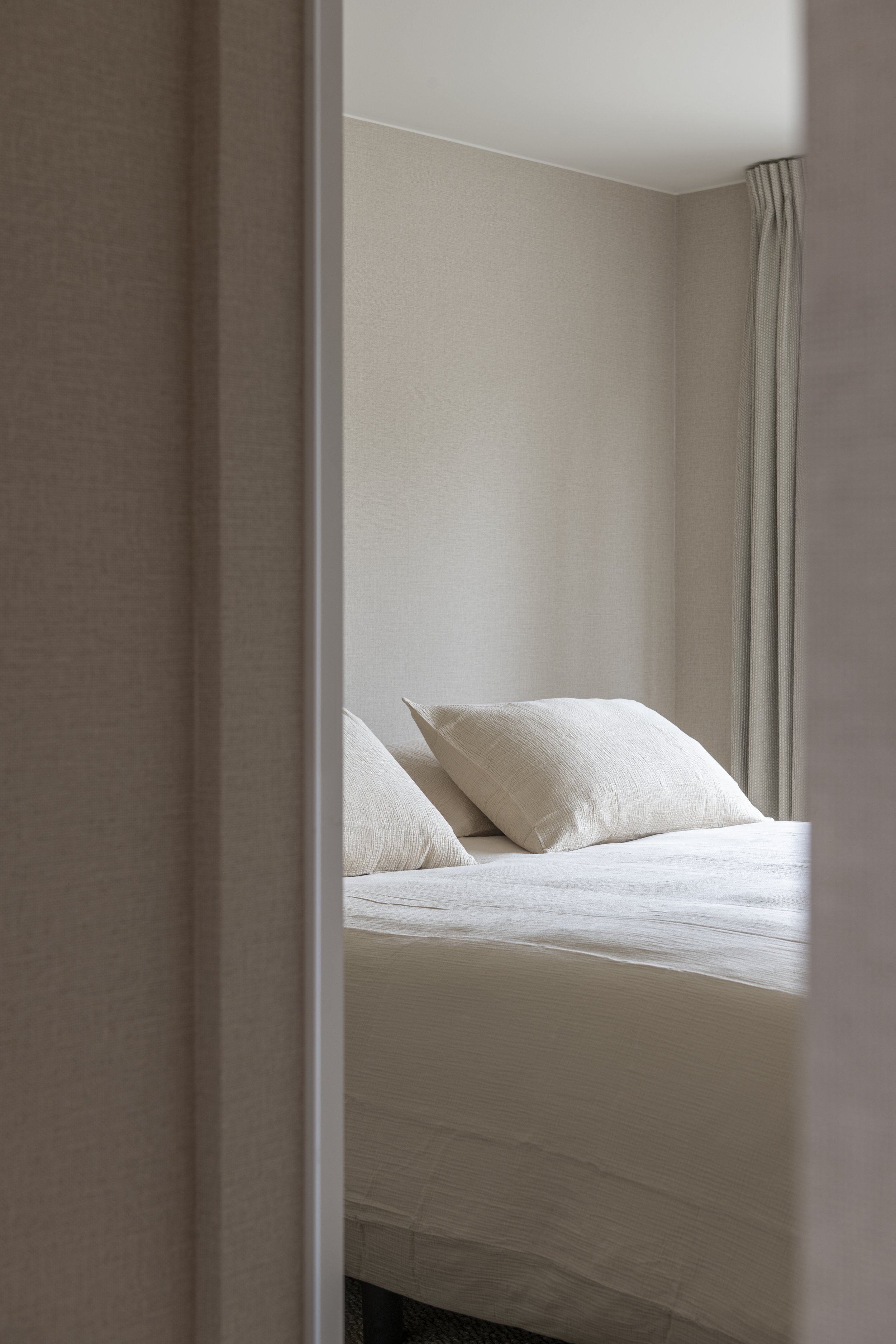 A hotel bedroom seen through an open door, featuring a bed with neutral-colored pillows and bedding, beige walls, and floor-length curtains.