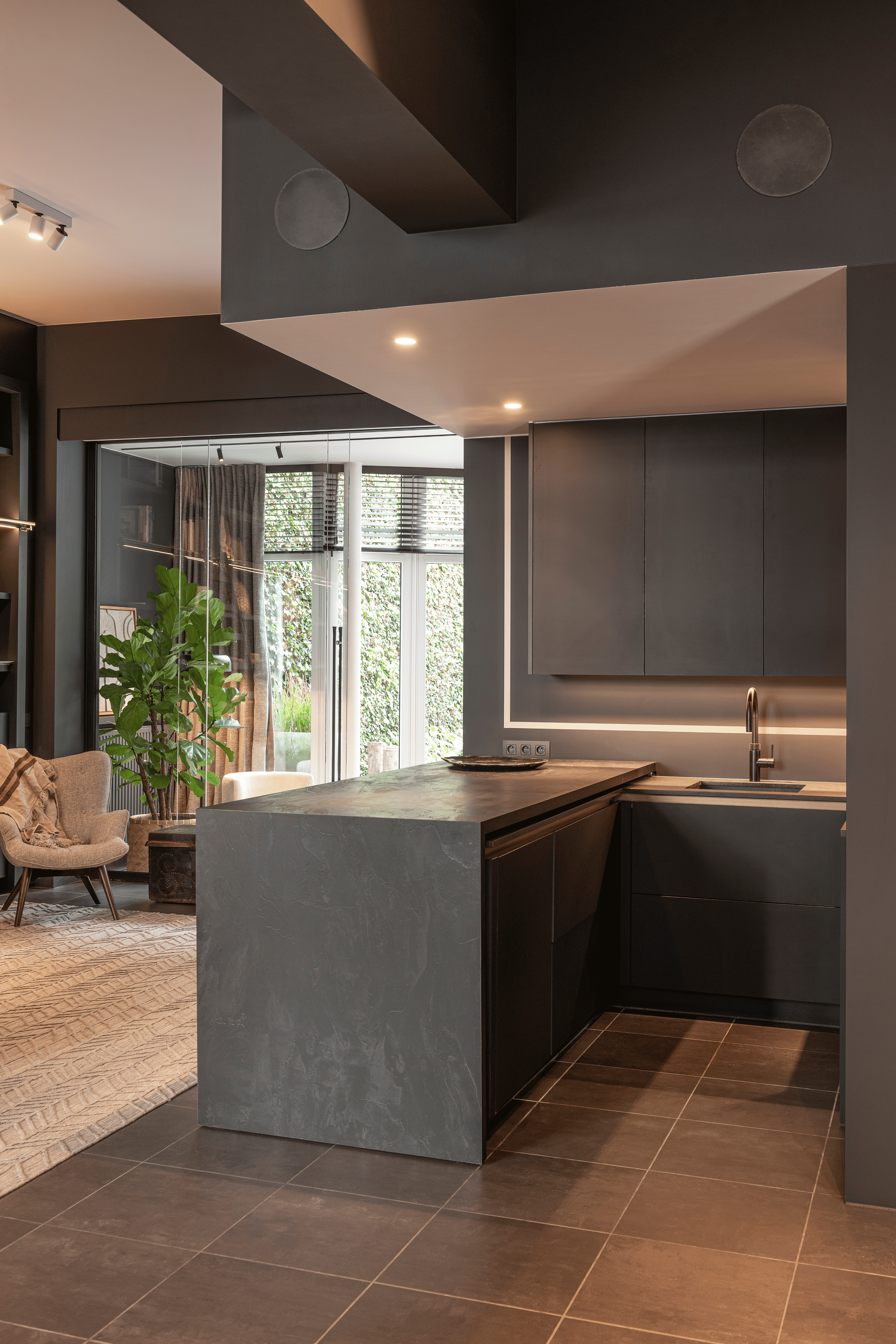 Modern kitchen with dark cabinetry, a marble countertop, and a view into a living area with large windows, curtains, and indoor plants.