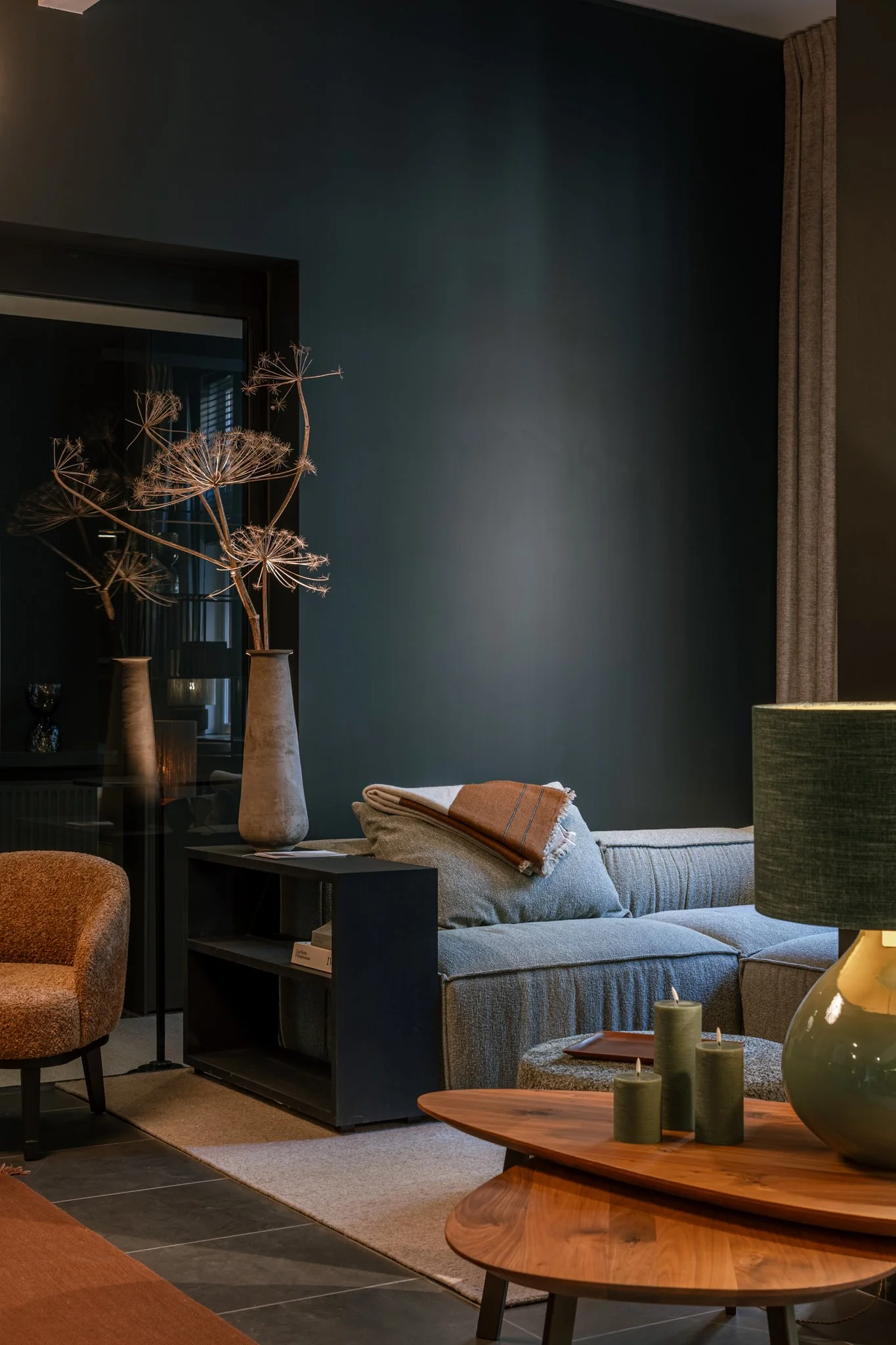 Living room interior with a beige sofa, a brown throw blanket, a black side table with decor, and a large wooden coffee table with candles and a decorative vase, against a dark blue wall and beige curtains.