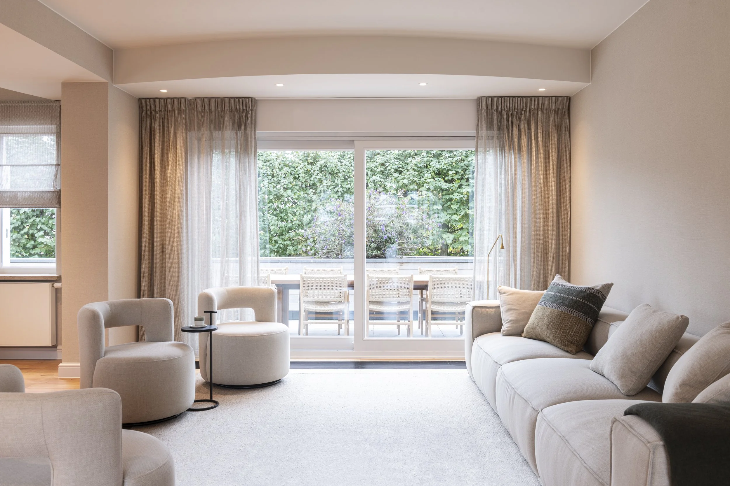Living room with white sofa, armchairs, and large sliding glass doors opening to a patio with chairs and greenery outside.