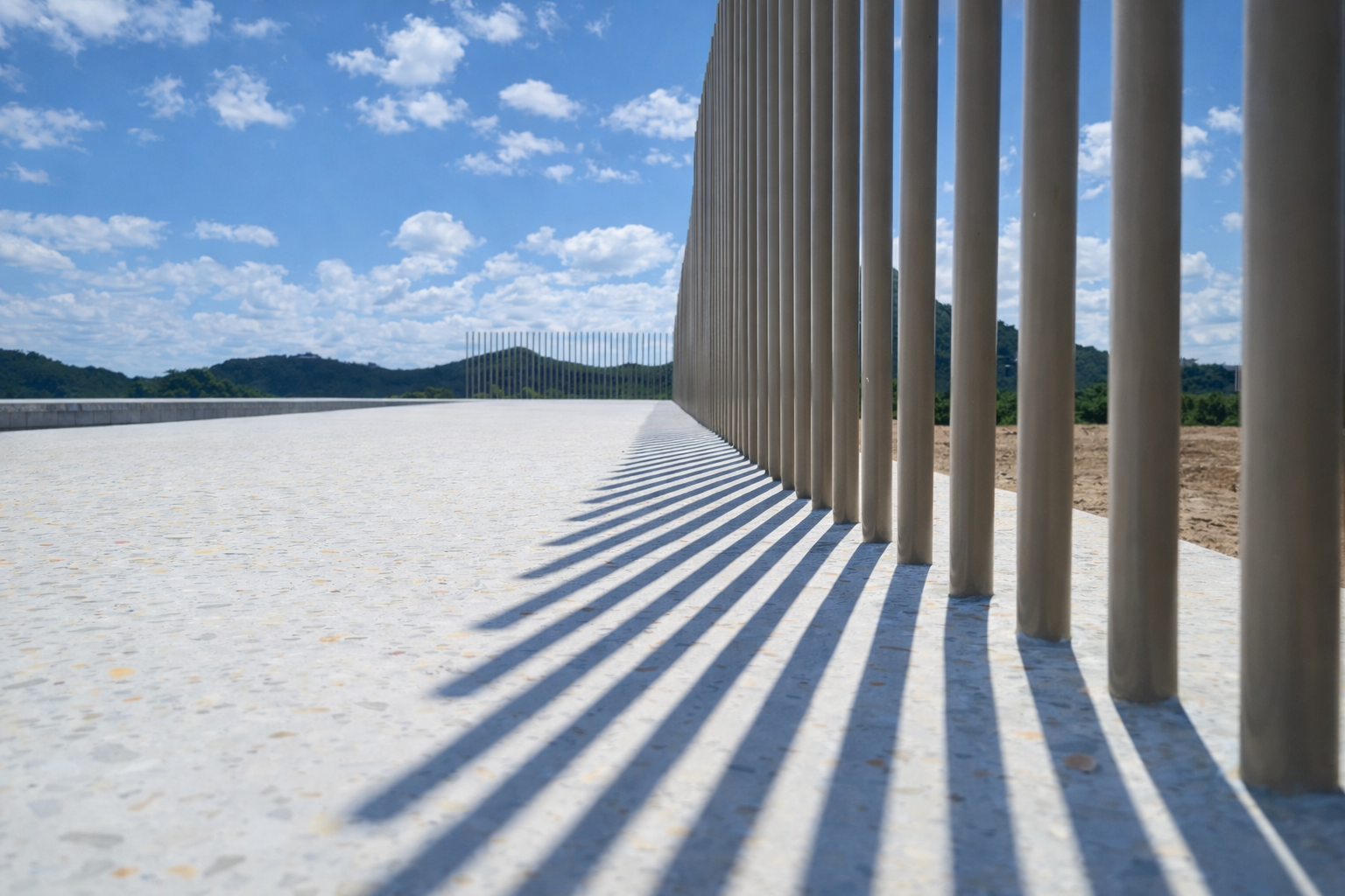 Metal fence with vertical bars casting elongated shadows on a white pavement against a blue sky with scattered clouds and distant green hills.