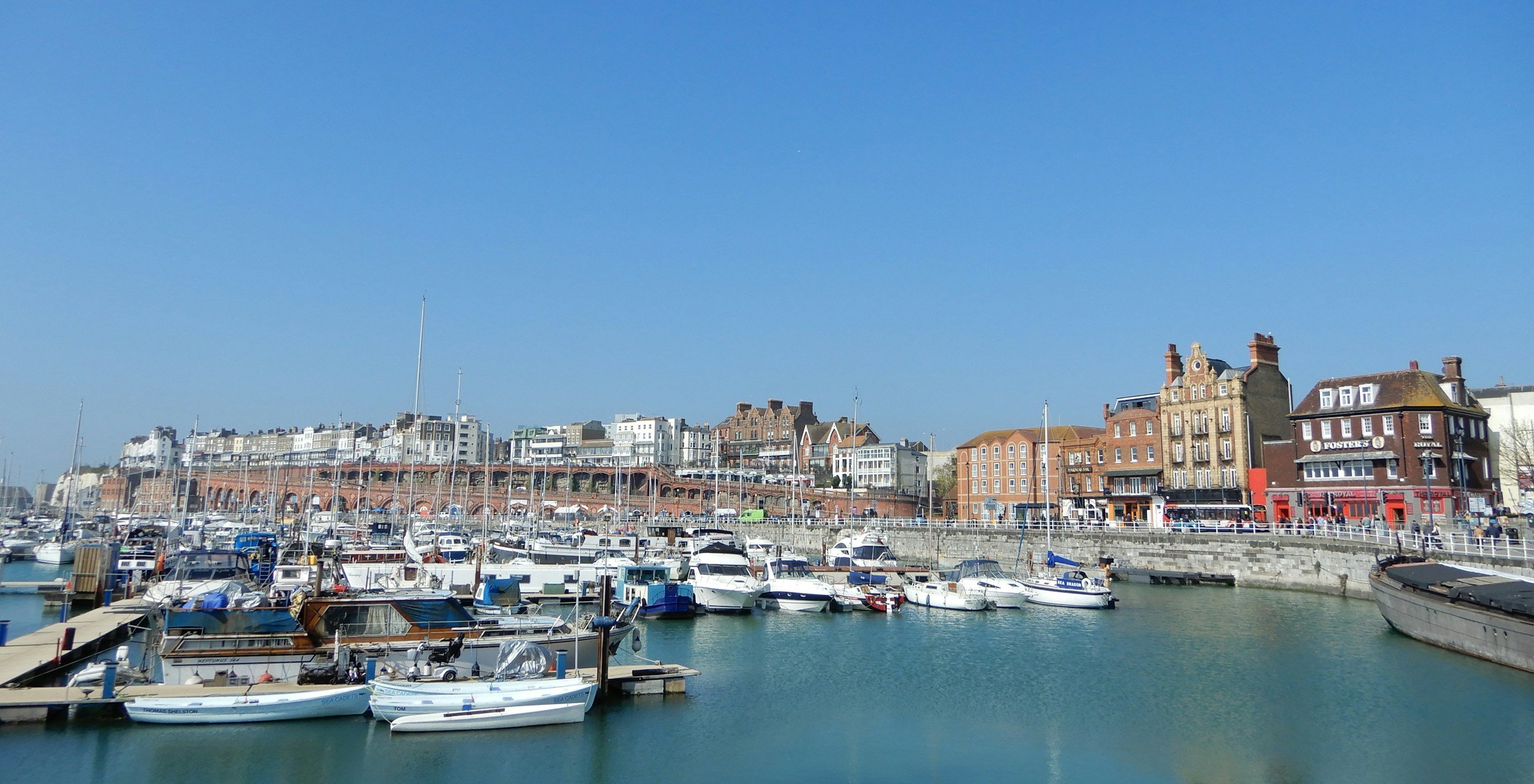 Harbor with boats docked in calm water, historic buildings along the shoreline, and a clear blue sky.