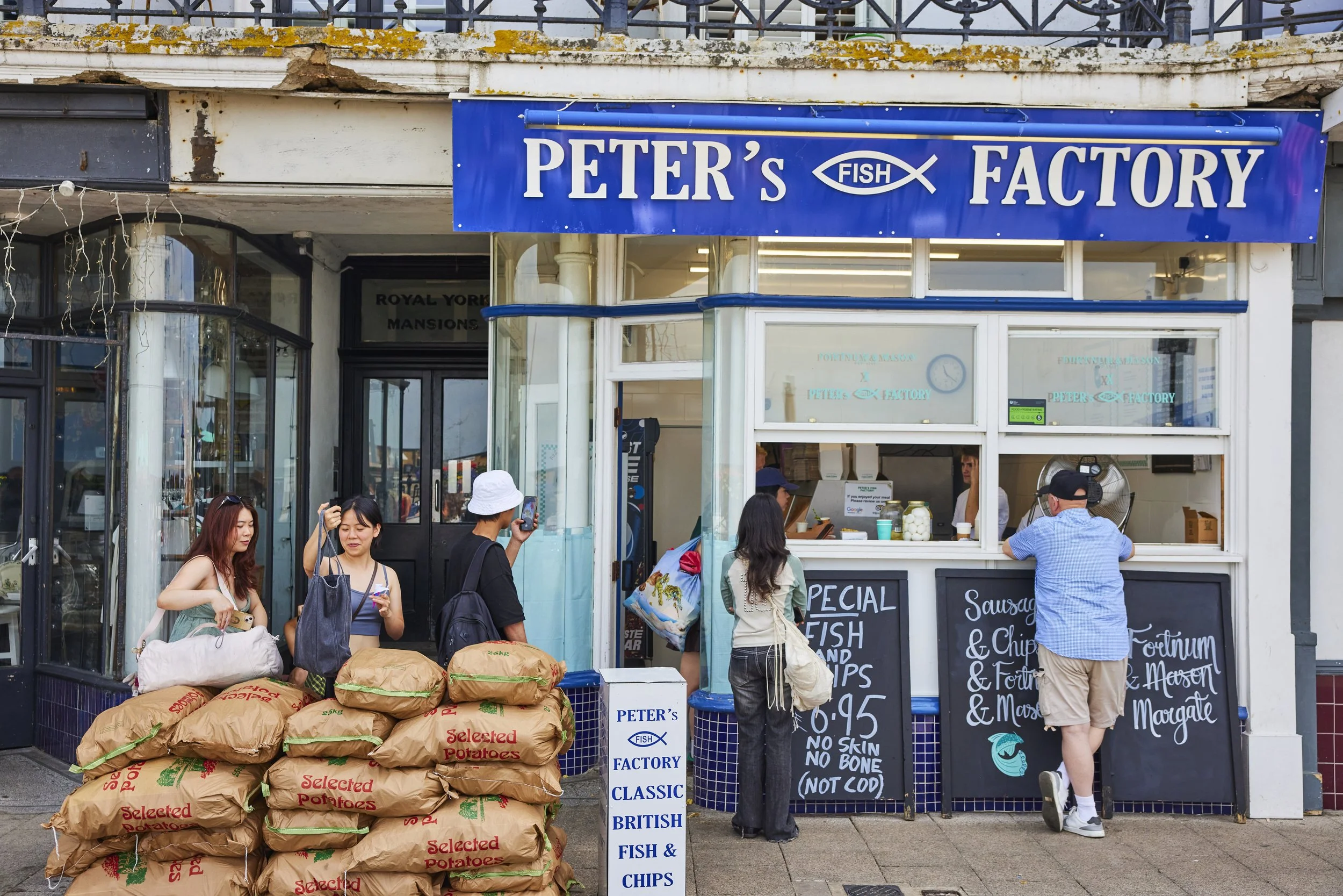 A fish shop named Peter's Fish Factory with people standing outside, some ordering and some waiting, and bags of potatoes on the sidewalk.