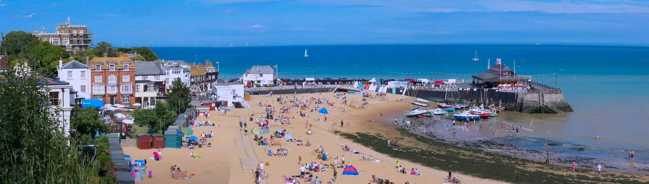 Beach scene with people relaxing on the sand, colorful umbrellas, boats docked near a pier, and a seaside town with houses and a restaurant in the background along the coast.