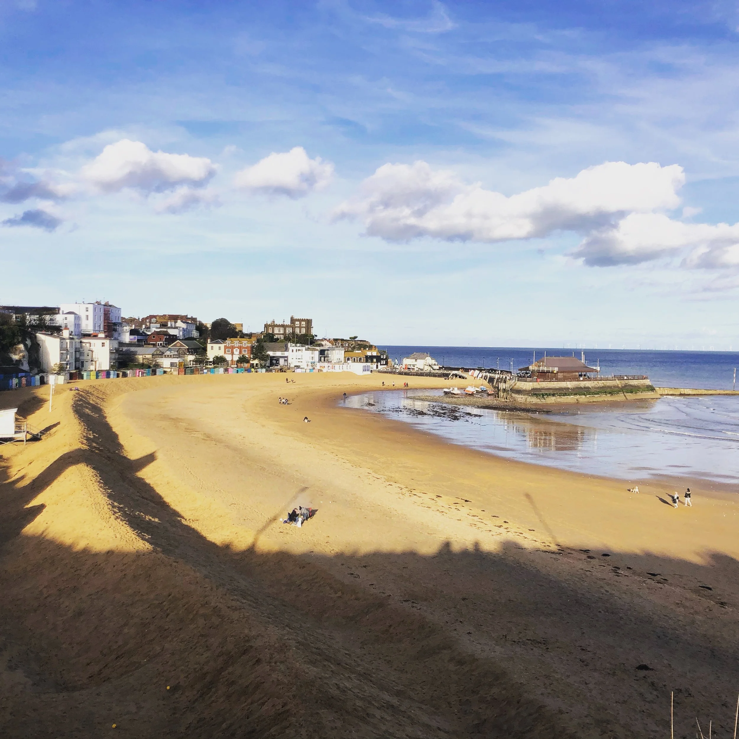 A quiet beach with golden sand, a few people sitting and walking, a small harbor with boats, and a row of colorful houses along the shoreline under a partly cloudy sky.