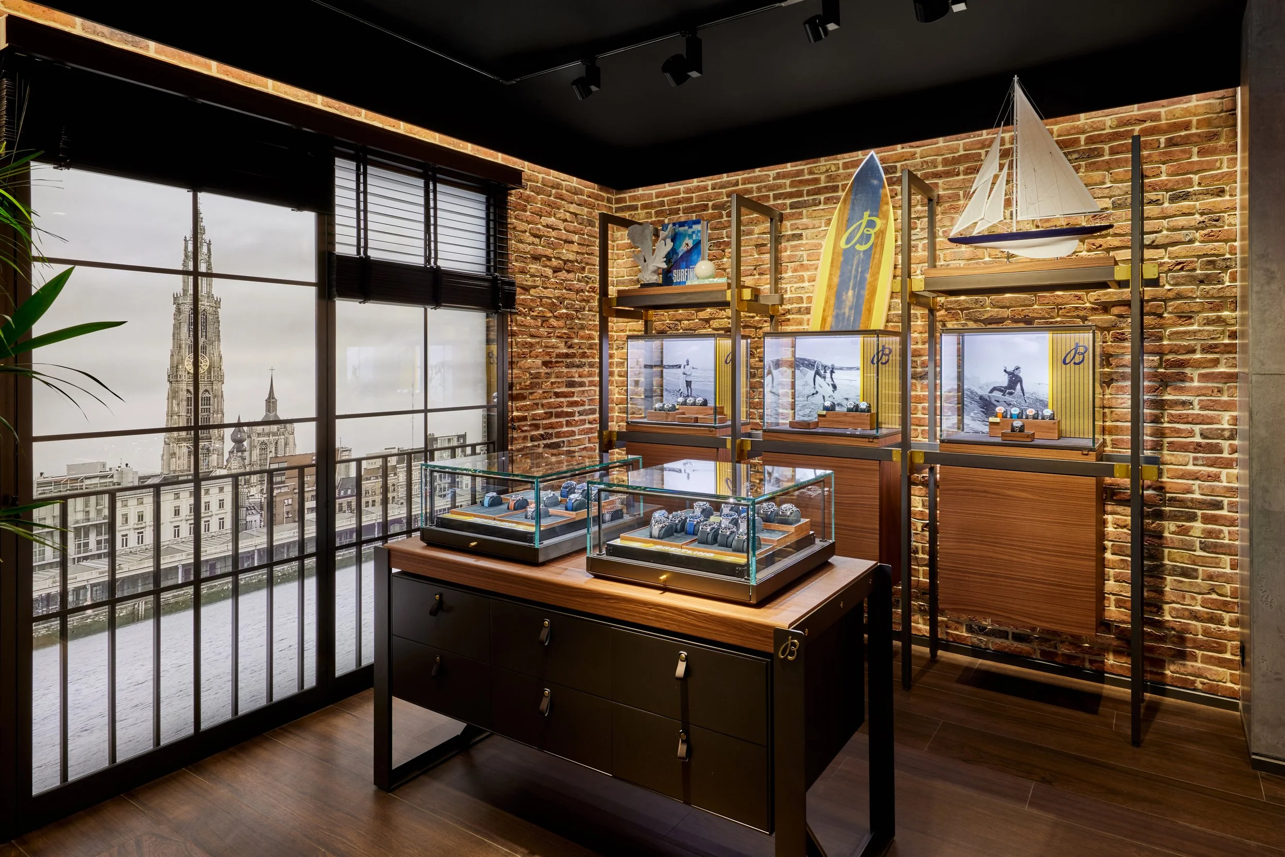 Interior of a jewelry store with display cases of watches, brick walls, large windows with a city skyline, and nautical decor including a model sailboat and surfboard.