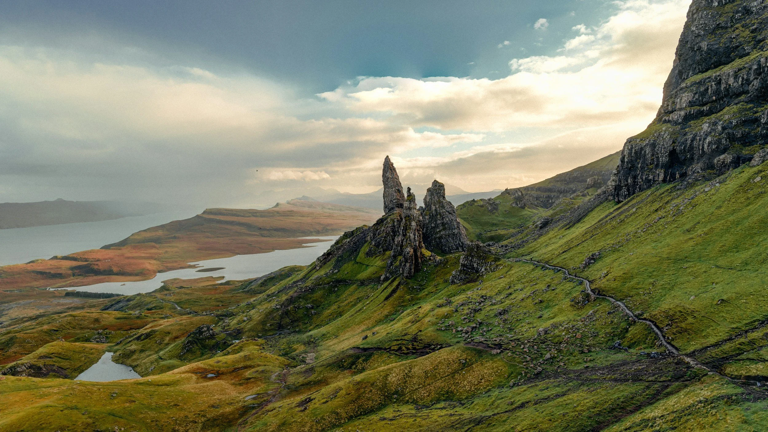 A scenic landscape of a green hillside with rocky formations and a body of water, under a sky with clouds.