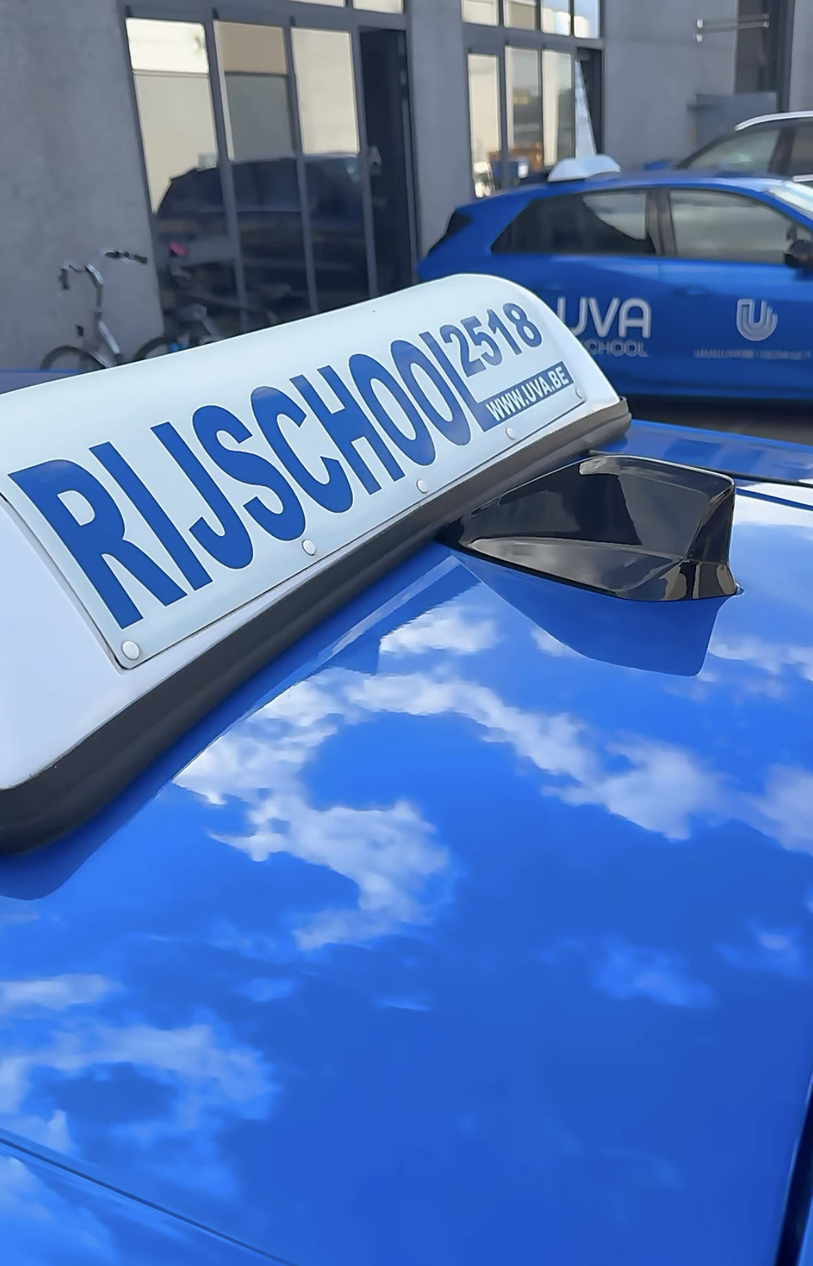 Close-up of a blue emergency vehicle with a sign reading 'Rijschool' and a website URL, parked outside a building with glass windows, reflecting the sky.