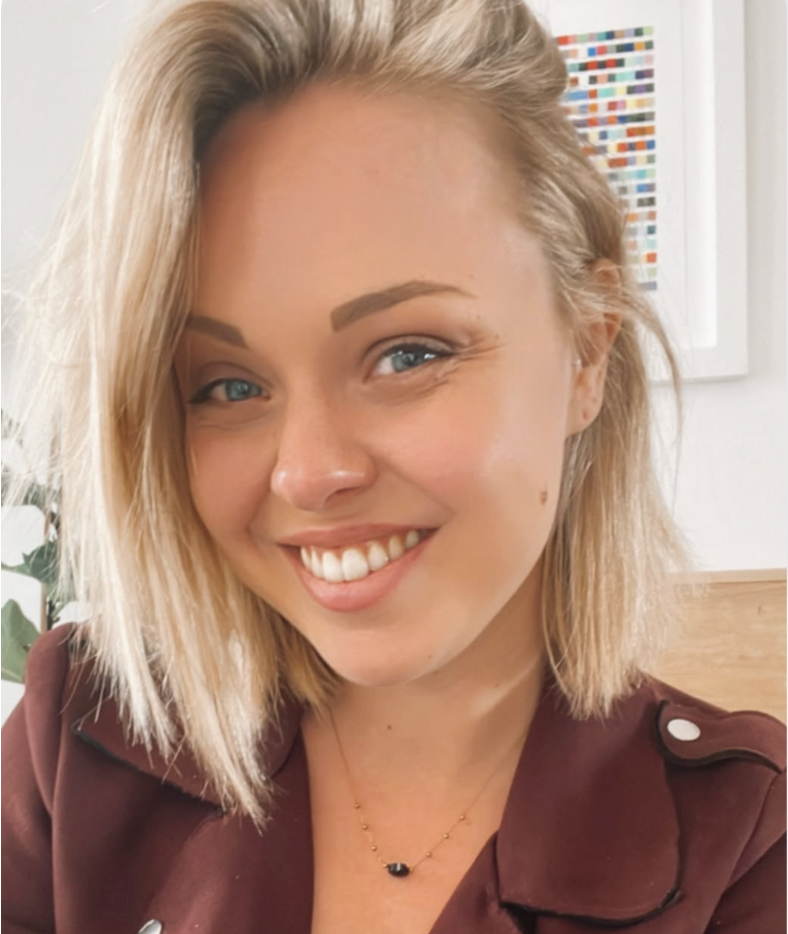 Close-up van een glimlachende vrouw met blond haar en blauwe ogen, gekleed in een bruine jas en een zwart-gouden ketting, in een goed verlichte kamer met kunstwerken op de achtergrond.