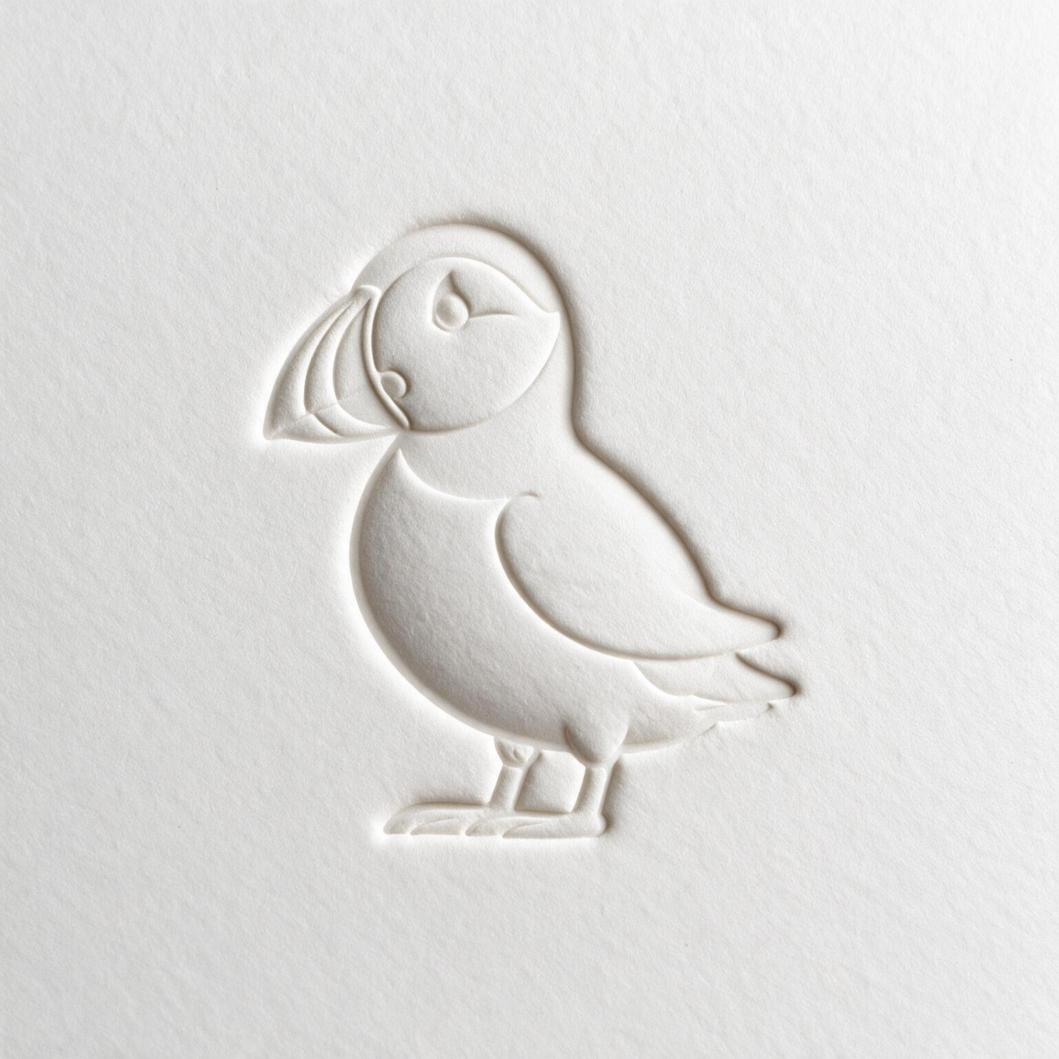 Embossed image of a puffin with a small eye, wing, tail, and standing legs on white textured paper.