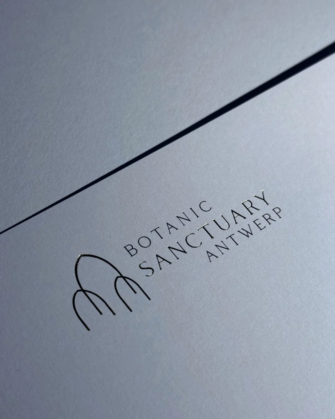Close-up of printed text on paper that reads 'Botanic Sanctuary Antwerp' with a minimalist graphic of three hills or arches.