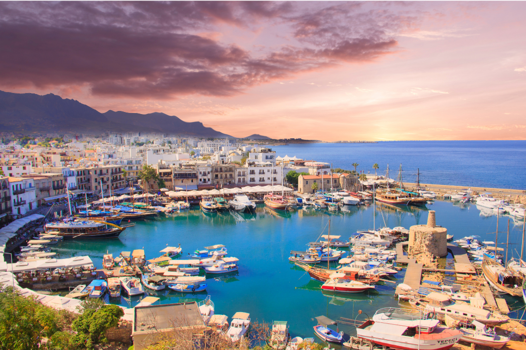 Scenic view of a harbor with boats docked, surrounded by white buildings, with mountains in the background and a sunset sky.