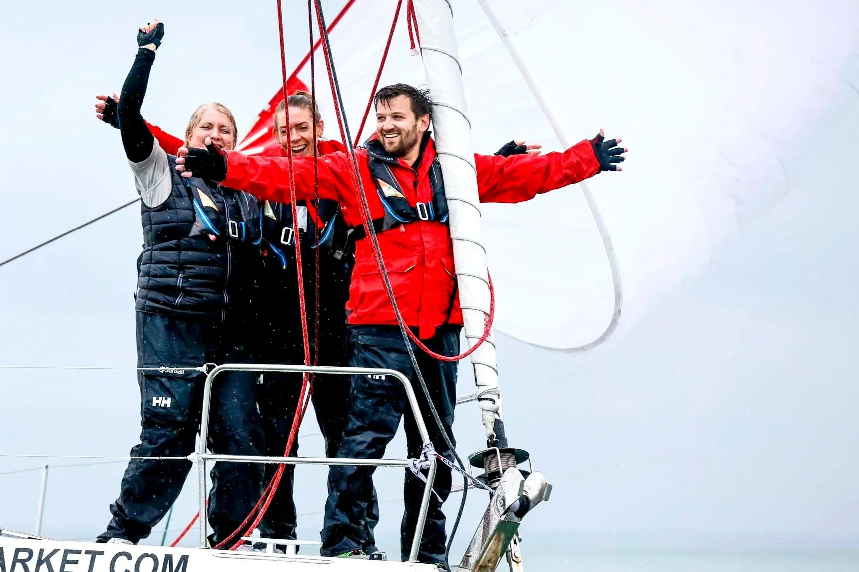 Three sailors celebrate on a sailboat with arms outstretched, wearing sailing gear and life jackets, with the sail partially visible behind them and a cloudy sky.