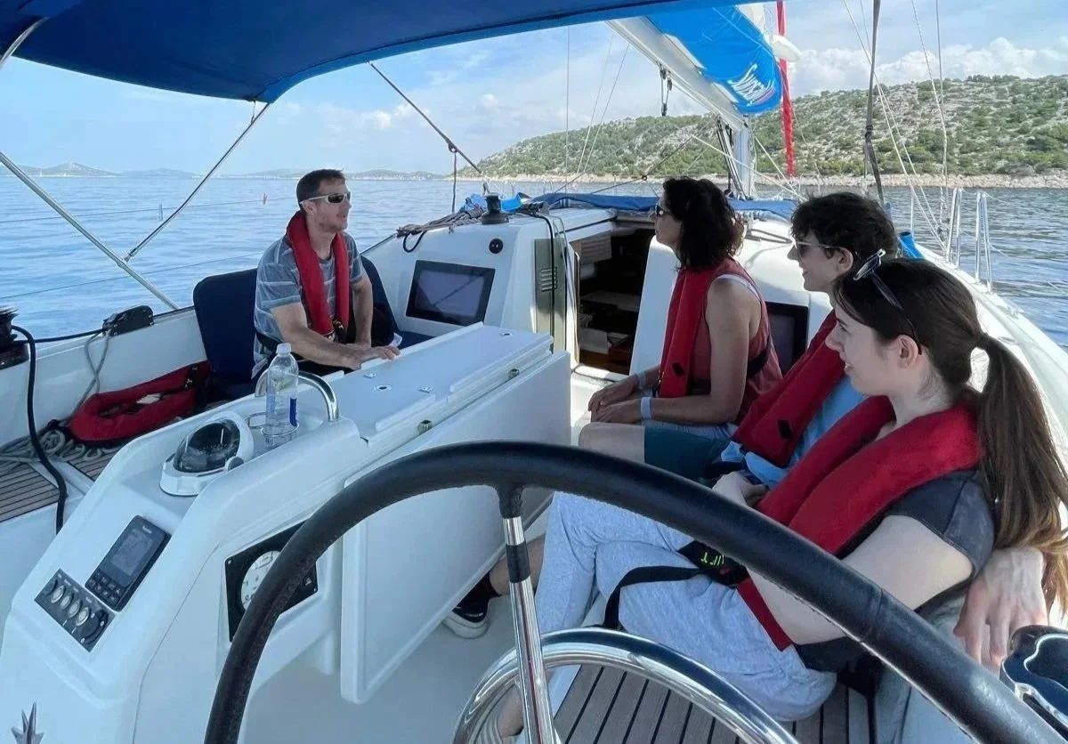 Four people sitting on a boat with life jackets, facing an instructor at the helm. The boat is on a calm body of water with land and hills in the background, under a partly cloudy sky.