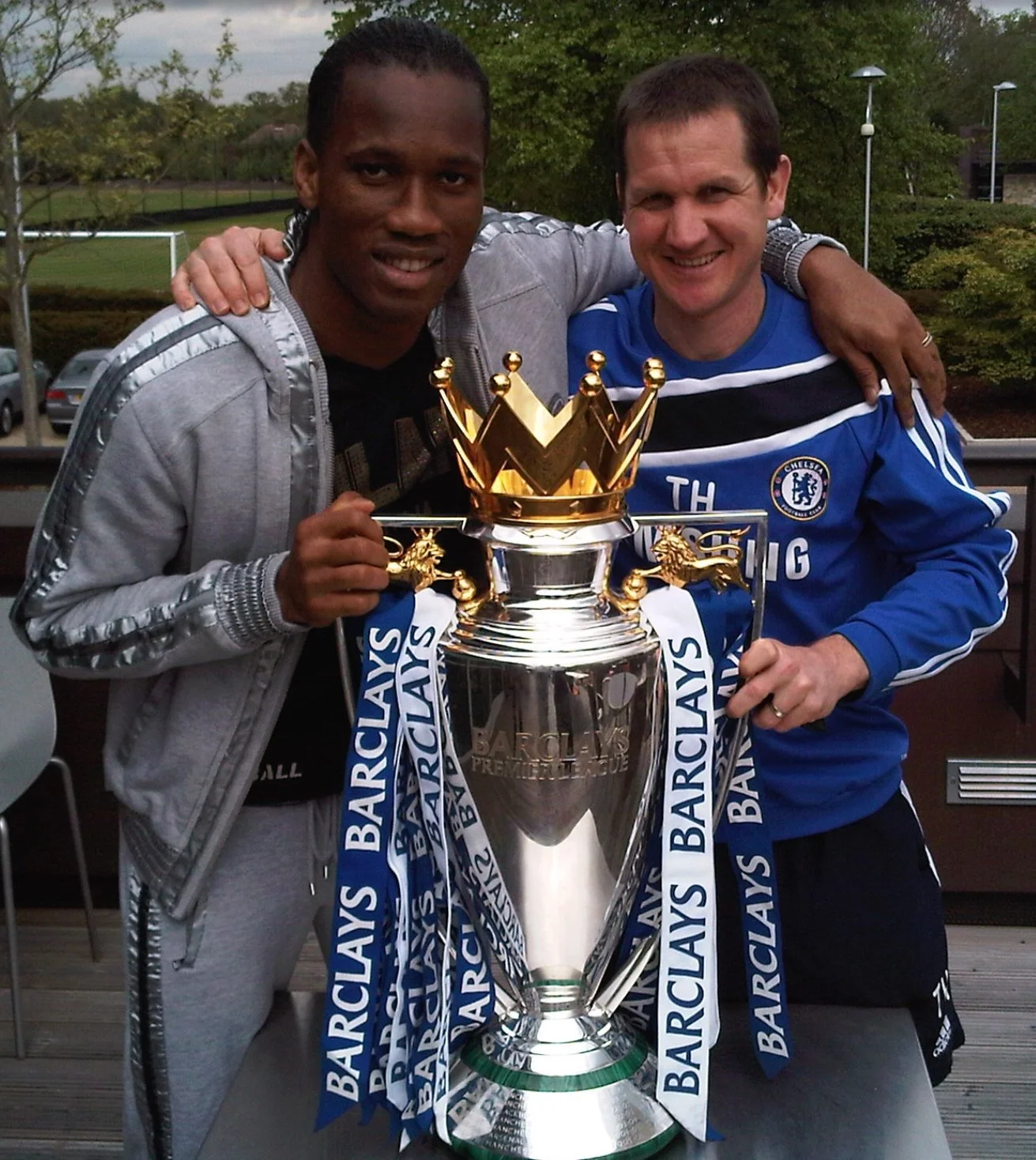 Two people holding the Premier League trophy, one wearing a Chelsea training top