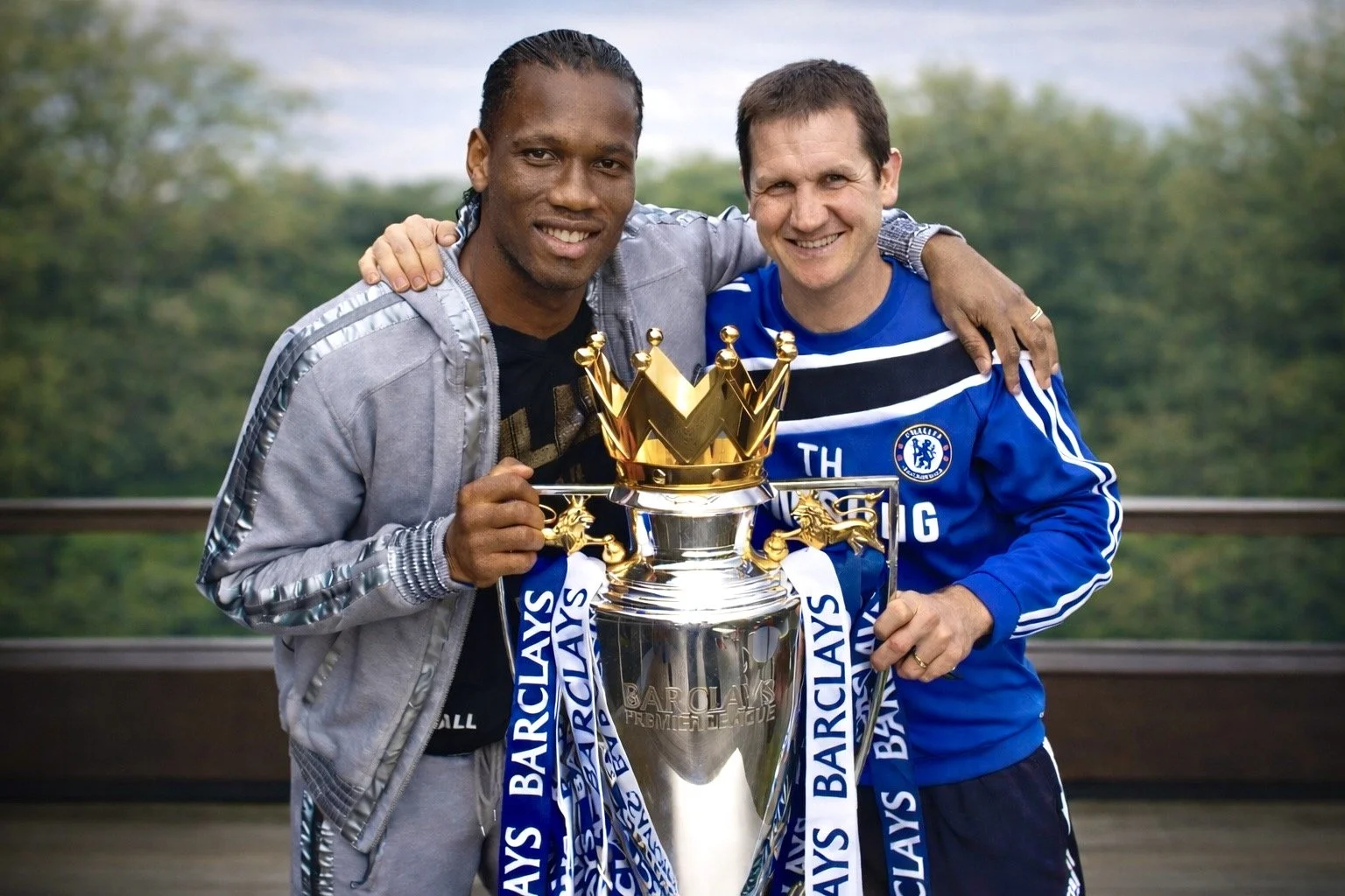Two men smiling, one in a gray jacket and the other in a Chelsea football shirt, holding a large Barclays Premier League trophy with blue and white ribbons, outdoors with a blurred green background.