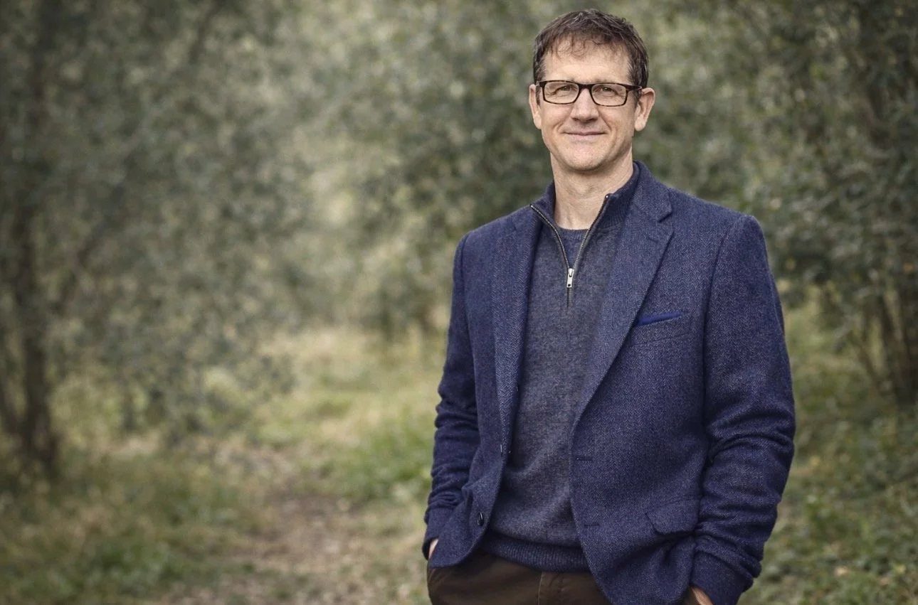 A man wearing glasses and a blue blazer standing outdoors on a trail surrounded by trees.