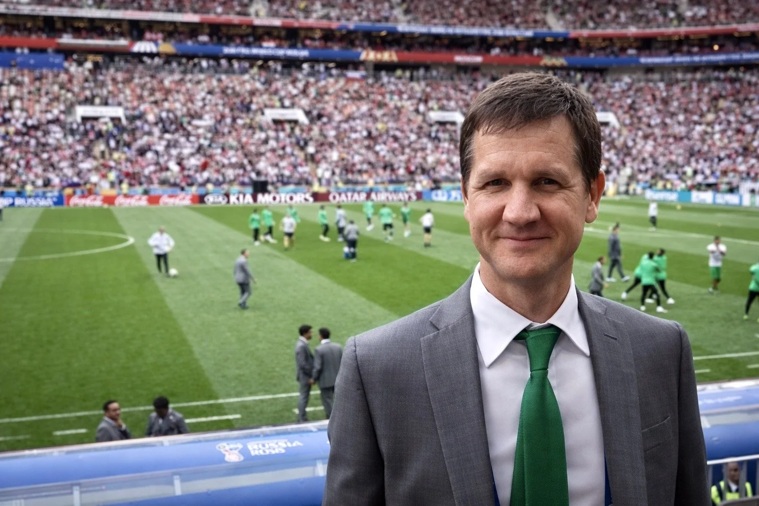 A man in a gray suit with a white shirt and green tie stands on the sidelines of a crowded football stadium, smiling with the field and players in the background.