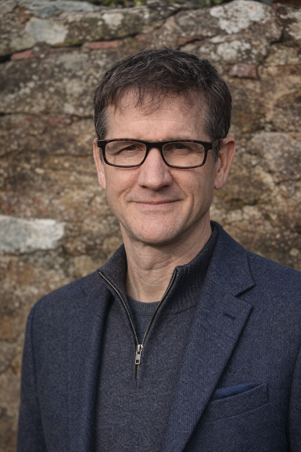 Portrait of a middle-aged man wearing glasses, a dark blazer, and a zip-up jacket, standing outdoors in front of a stone wall.