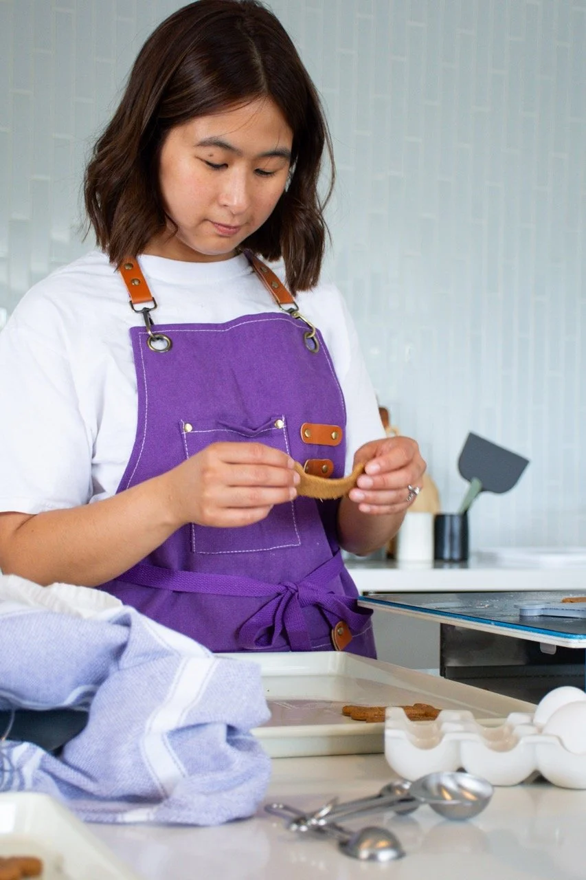 A woman with shoulder-length brown hair, wearing a purple apron over a white shirt, is wrapping a cookie in a brown paper strip in a kitchen setting.