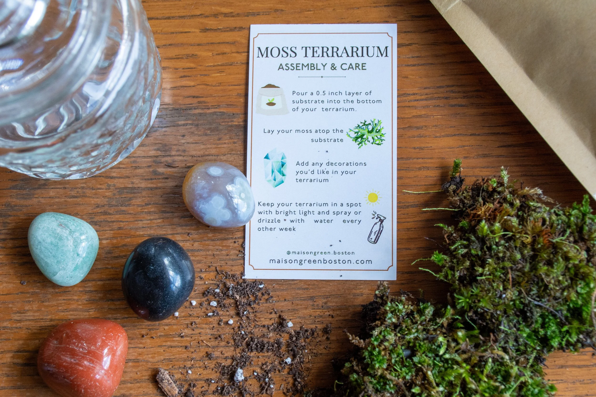 Instructions for moss terrarium assembly on a wooden surface, with colored stones, moss, a jar, and a potting soil bag.