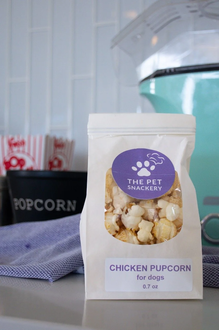 A white bag of chicken popcorn treats for dogs from The Pet Snackery, with a purple label featuring a paw print and bone graphic. The bag is placed on a surface with popcorn and other snack packages in the background.